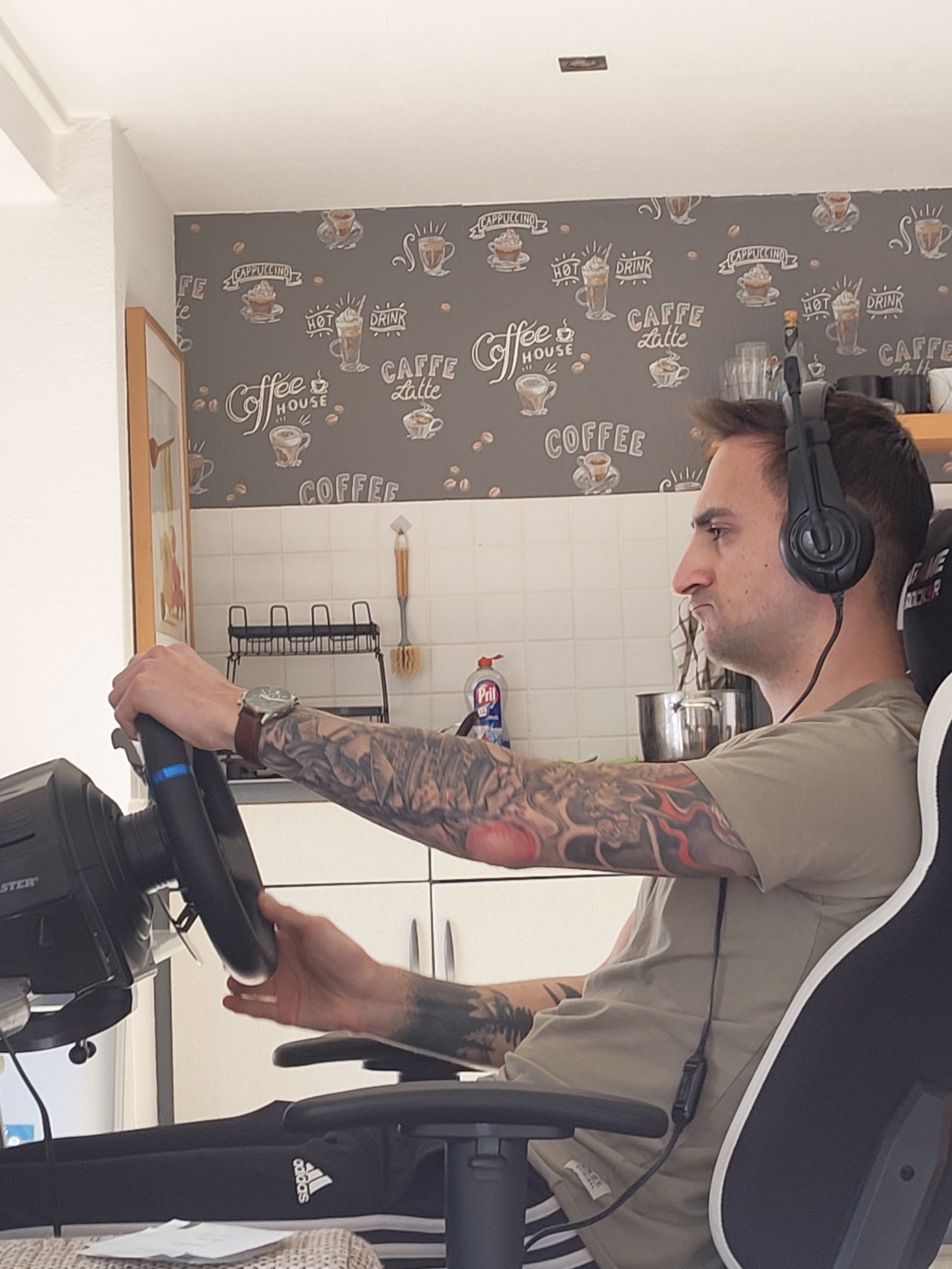 An adult male is observed in a domestic interior in Leidersbach, Germany, seated in a black and white gaming chair, actively engaged with a racing simulator. He is holding a black Thrustmaster steering wheel with both hands. The man has short, dark hair, stubble, and wears a plain olive-green t-shirt, dark Adidas track pants with white stripes, and black over-ear headphones with a cable running down his chest. His left forearm and upper arm are heavily tattooed with a multicolored sleeve featuring what appears to be Japanese-style imagery, and a wristwatch with a brown strap is on his left wrist. The background features a kitchen area with a white tiled backsplash. Above the tiles, the wall is covered with dark grey wallpaper patterned with coffee-related illustrations and text. Visible on the countertop are a black two-tiered dish drying rack, a cleaning brush hanging on the wall, a blue bottle of Pril dish soap, stacked clear glasses, and a large metal pot. A white ceiling with a single rectangular light fixture is visible at the top.