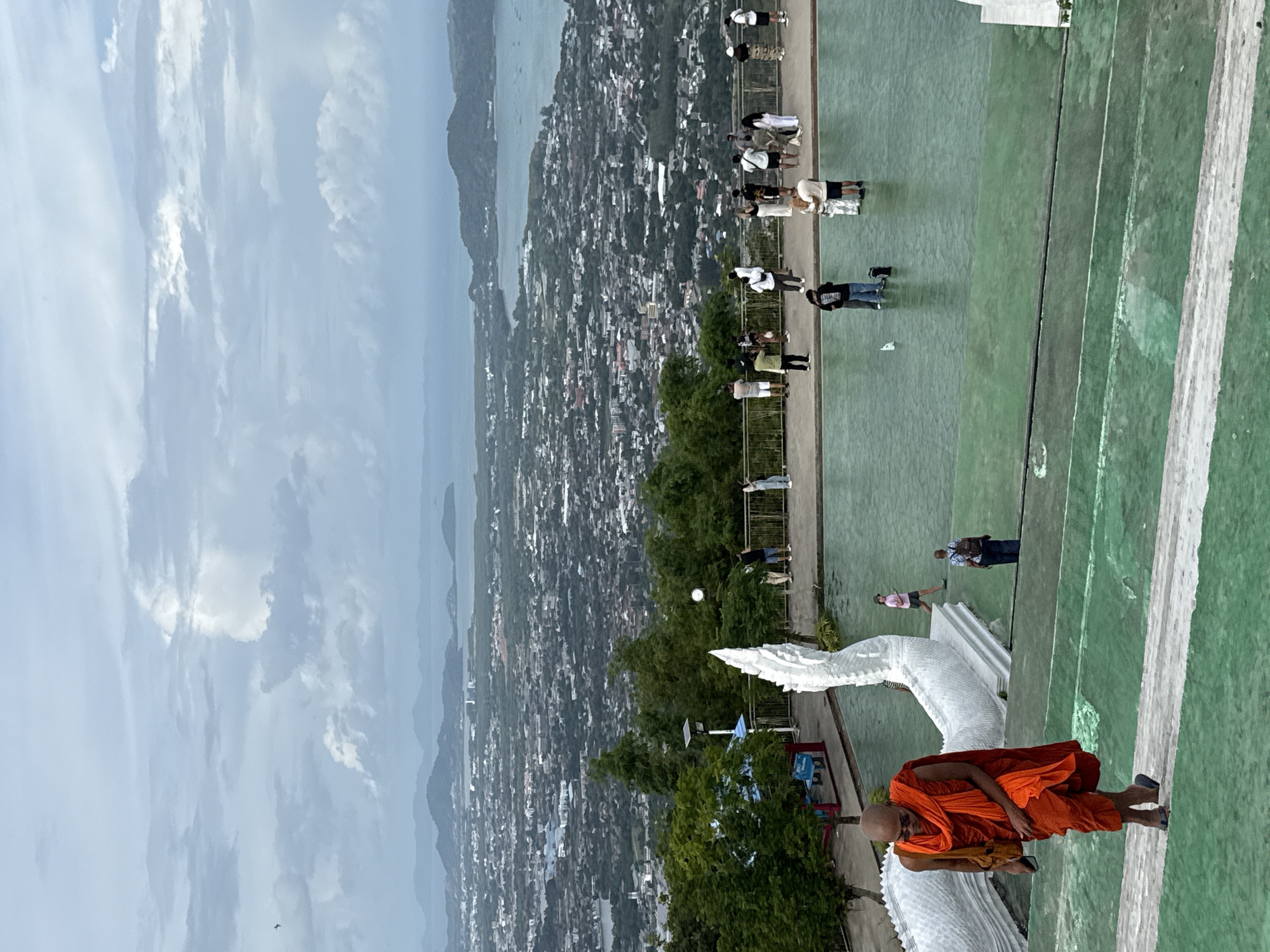 The image shows a panoramic view from a high vantage point overlooking the city of Karon, Thailand. In the foreground, a Buddhist monk in orange robes walks down a green-tiled stairway. Behind him, a large white serpent sculpture, likely a Naga, is visible. Further back, a group of tourists is gathered on a balcony, looking out at the city and the ocean. The city of Karon stretches out below, a dense collection of buildings with a bay and distant islands visible in the background. The sky is overcast with dramatic clouds. The overall scene suggests a popular tourist destination offering scenic views.