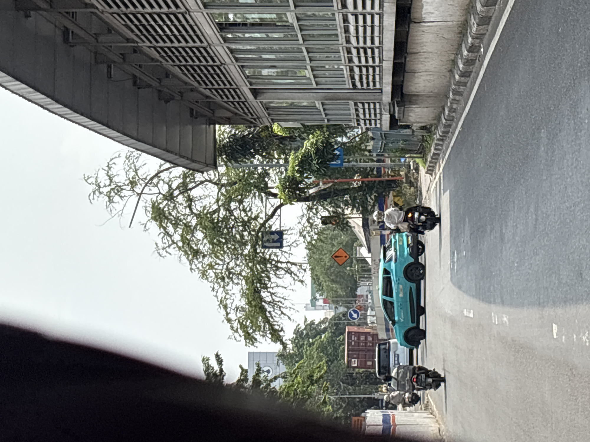 The image captures a street scene in North Jakarta, Indonesia. A light blue car is traveling on a gray asphalt road, accompanied by several motorcycles. The road is bordered by a concrete curb and a sidewalk. Overhead is a metal structure, likely a pedestrian bridge or part of a building. A tree with sparse foliage stretches across the frame, partially obscuring traffic signs. In the distance, buildings and trees are visible. The image is taken from within a vehicle, indicated by a dark section at the bottom of the frame. The sky is overcast.