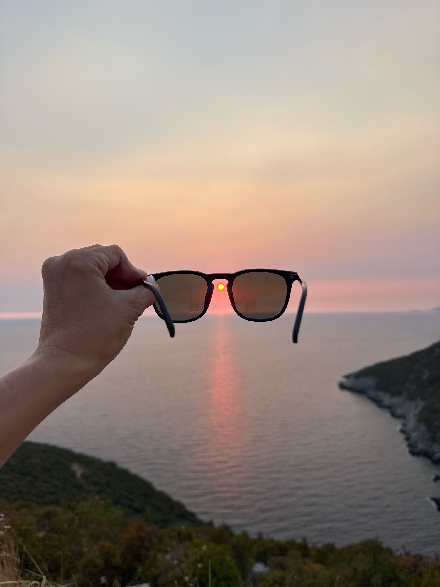 A hand holds a pair of black-framed sunglasses in the foreground, obscuring part of the view of a sunset over a body of water. The sun, appearing as a bright orange orb, is visible through the right lens of the sunglasses, casting a pinkish reflection on the water. The sunglasses are angled downwards. In the background, the sea stretches out to the horizon, meeting a hazy, pastel-colored sky that transitions from pale blue at the top to pink and orange near the horizon. To the right, a rocky, tree-covered coastline juts into the water. The overall scene suggests a coastal location, possibly near Zagora, Montenegro, during the evening.