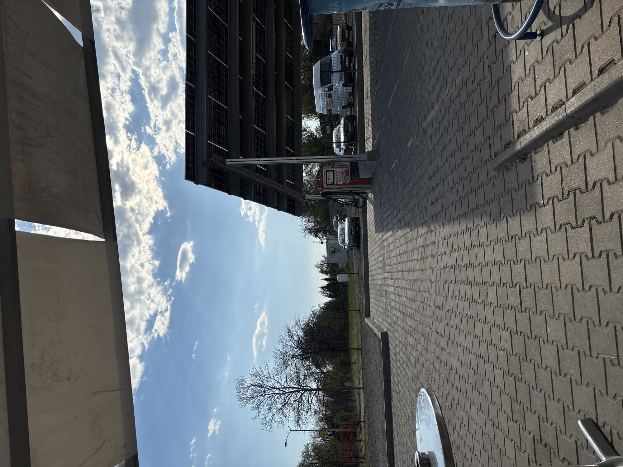 The image depicts a parking area in Chirpan, Bulgaria. The foreground shows interlocking concrete pavers extending into the distance. Various vehicles, including cars and a white van, are parked under a solar panel structure that provides shade. A pay station for car washing or charging is also visible. Trees are visible in the background, along with a clear blue sky with scattered clouds. The image is taken from a low angle, and some objects in the foreground are partially obscured by a structure with a metal frame and a fabric canopy.