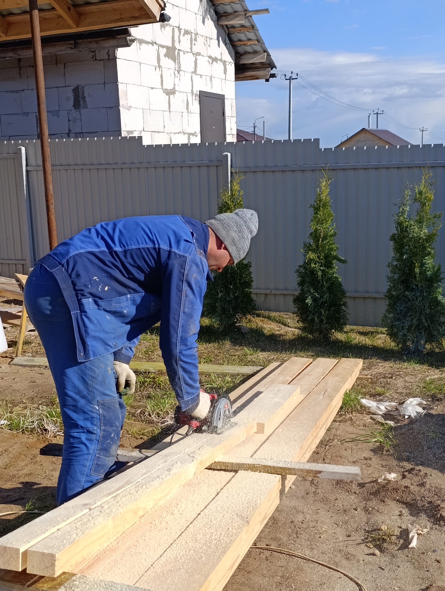 A man is captured mid-action, cutting a wooden plank with a circular saw in what appears to be a yard or construction site in Chaikovsky, Russia. He is dressed in blue work overalls and a grey beanie, with work gloves visible on his hands. Sawdust covers the wooden planks and the surrounding ground, indicating active work.
The scene unfolds outdoors on a bright, sunny day, with the sun casting clear shadows. In the background, a light grey corrugated metal fence runs horizontally across the image. Behind the fence, three slender evergreen trees, possibly thuja, are planted. To the left, an unfinished building constructed from white aerated concrete blocks can be seen, hinting at ongoing construction. Further in the distance, above the fence line, other houses with brown roofs and power lines stretching across a partially cloudy blue sky are visible, suggesting a suburban or semi-rural setting. The ground beneath the man's feet is a mix of dirt and sparse grass, with some scattered debris, including what looks like white plastic bags. No visible text is present in the image.