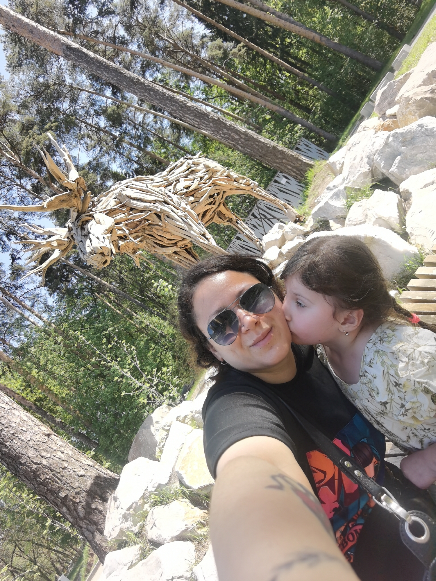 The image is a selfie of a woman with short dark hair and sunglasses, being kissed on the cheek by a young girl with dark hair in a braid. They are outdoors in a park or wooded area. Behind them is a large sculpture of an animal, seemingly made of driftwood. Trees and other greenery are visible in the background. The location is within the city of Perm, Russia.