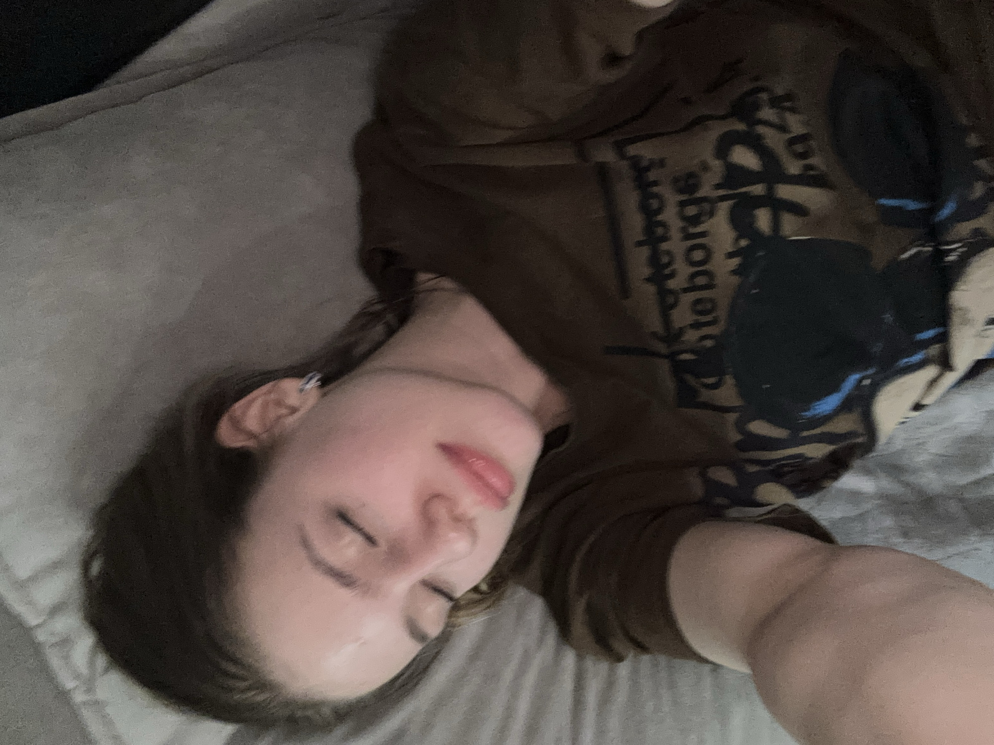 A close-up, high-angle, slightly blurry photograph shows a young woman lying down with her eyes closed. She has brown hair pulled back from her face. She is wearing a dark brown t-shirt with a graphic print that includes text that appears to read "Göteborgs". The woman is lying on a grey, textured surface, likely a bed or sofa. The lighting is dim, casting a soft glow on her face. There is a hint of pink on her lips and cheeks, possibly from makeup or natural flush. She is wearing a small hoop earring in her visible ear. The overall impression is that she is resting or sleeping. The image is a selfie, as indicated by the angle and the woman's arm reaching into the frame. The location is not specified in Rakhya, Russia.