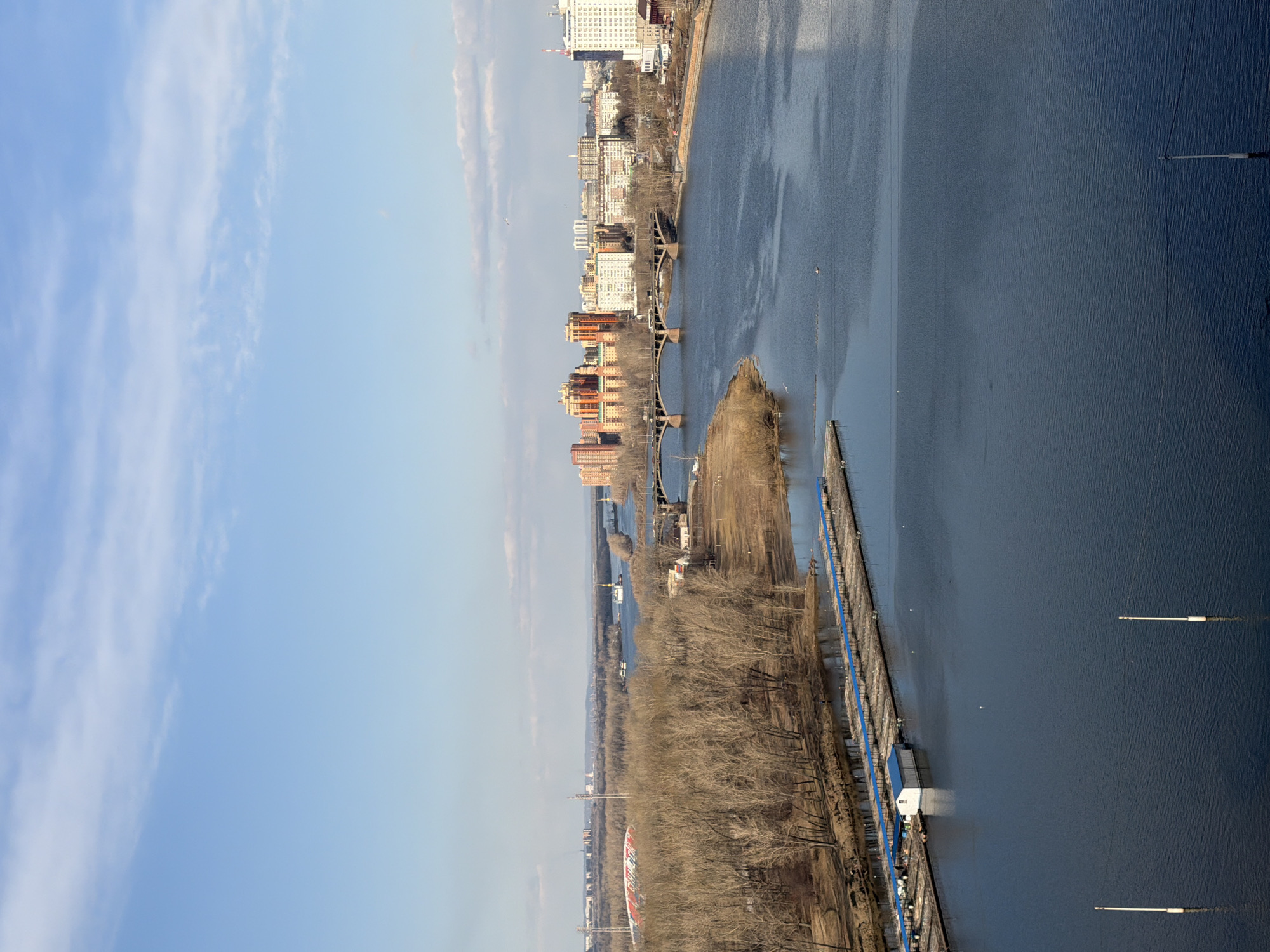 The image depicts a view of the Yenisey River in Krasnoyarsk, Russia. The river runs vertically through the frame, dividing the urban landscape. On the left bank, a line of leafless trees stretches alongside a road. Further back, various buildings and city infrastructure are visible. Multiple bridges span the river, connecting the two sides. In the foreground, a long, narrow island sits in the river. The right bank of the river shows the continuation of the urban area with additional buildings and structures. The sky is blue with scattered clouds. Some people are on the docks by the water.