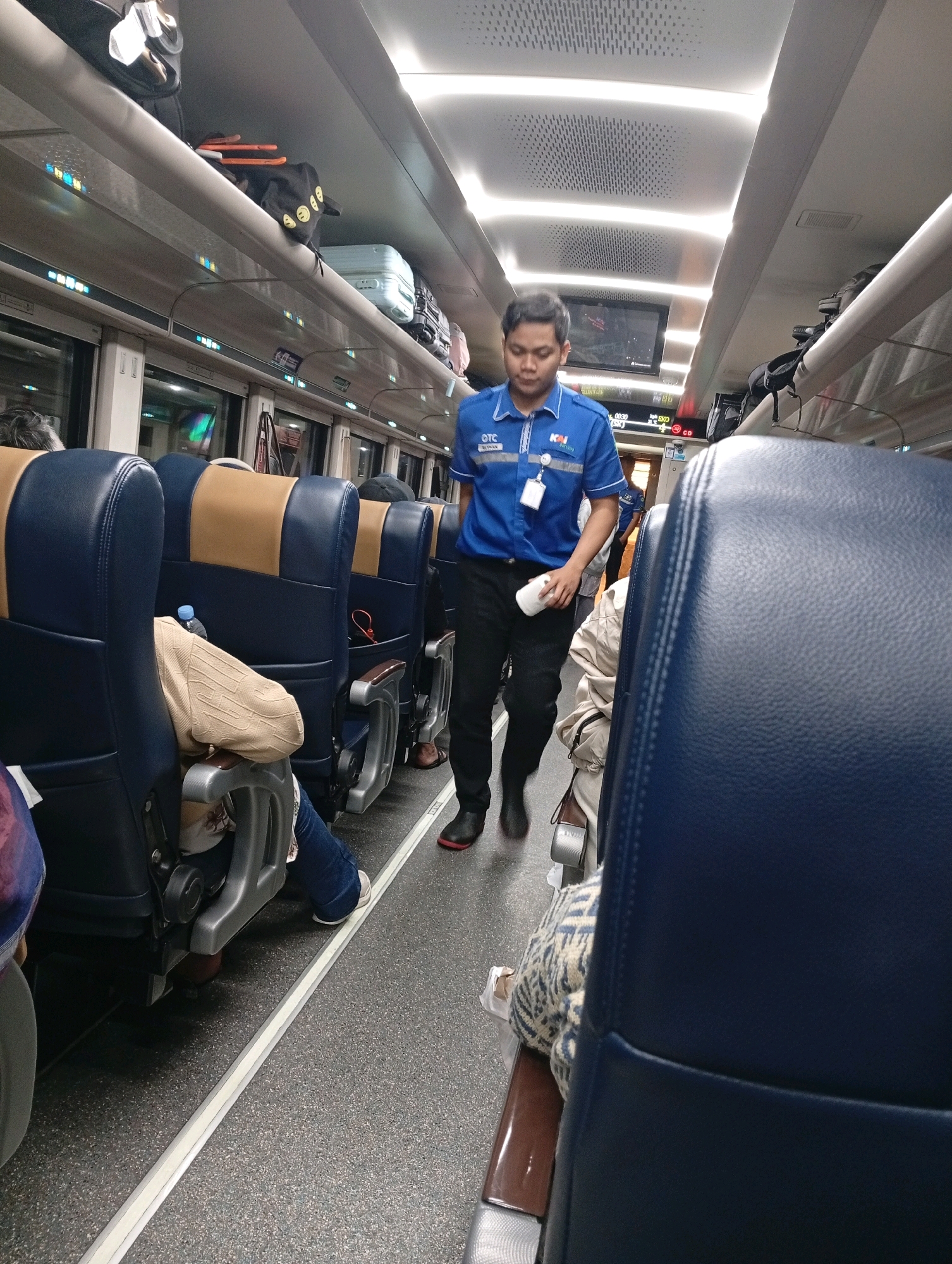 A man wearing a blue uniform with "KAI" and "OTC" logos is walking down the aisle of a train. He is holding a white cup or container in his right hand. The train car has rows of blue and tan seats, some occupied by passengers. Luggage is stored on overhead racks. The floor is grey and speckled, with a white strip running along the aisle. The image was taken inside a train carriage, possibly during a journey to or from Tegal, Indonesia, as suggested by the context.