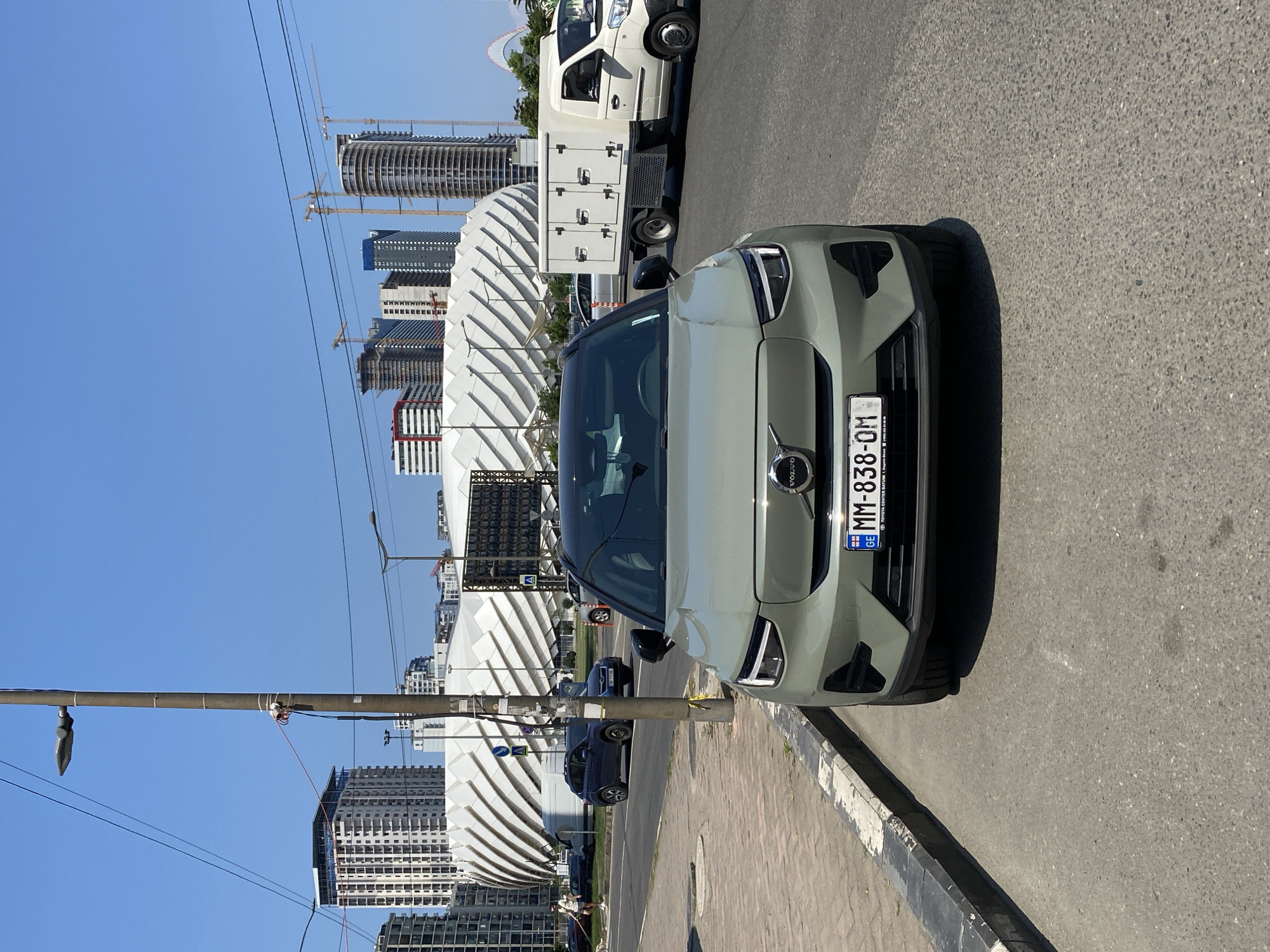 An exterior, eye-level, full frontal shot captures a light green Volvo XC40 electric SUV parked on the side of a street in Batumi, Georgia, on a clear, sunny day. The car's license plate reads "MM-838-OM". In the background, several modern buildings are under construction, with cranes visible. A distinctive white, wave-like architectural structure, possibly a stadium or concert hall, dominates the mid-ground. A white box truck is parked on the street to the right of the Volvo. On the left side of the image, a person is walking a dog on the sidewalk. The street is paved with asphalt, and a concrete sidewalk with a curb is visible in the foreground. A lamppost with wires is positioned near the parked car.