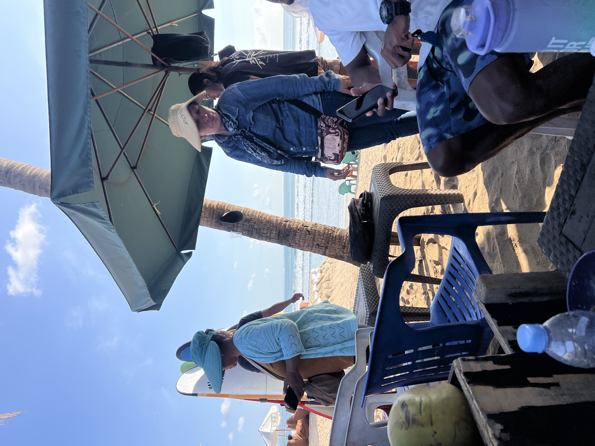This image captures a scene on a beach in Kuta, Indonesia. In the foreground, a person wearing blue patterned swim shorts and a white t-shirt is seated, holding a mobile phone. To the left, a partially visible coconut sits in a wooden crate. A blue plastic chair is positioned in front of the crate. Further back, a woman wearing a blue cardigan and a blue visor stands on the sand, holding a smartphone. She is near surfboards leaning against a palm tree. To her right, another woman in a denim shirt and a straw hat stands under a large green umbrella. A second person, wearing a black t-shirt with white text and tan shorts, stands behind her. The background features the ocean with gentle waves and a sandy beach with scattered chairs and umbrellas. The sky is clear and blue with a few clouds.