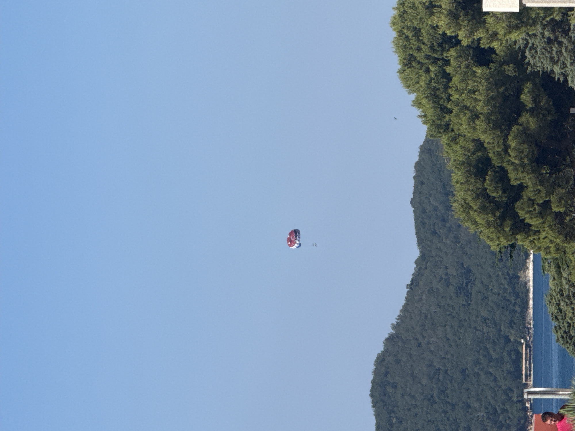 This media file depicts a parasailer in mid-air against a clear blue sky. The parasail is red and white, with a person attached below. The scene is set in a coastal area, with lush green trees covering a hillside in the background. Below the trees, a body of blue water, likely the sea, is visible. The image suggests a sunny day, possibly late morning or early afternoon, due to the bright lighting. The location cues of a coastline and abundant vegetation are consistent with Rafailovići, Montenegro. There is no visible text in the image. The overall impression is one of leisure and outdoor recreation in a scenic environment.