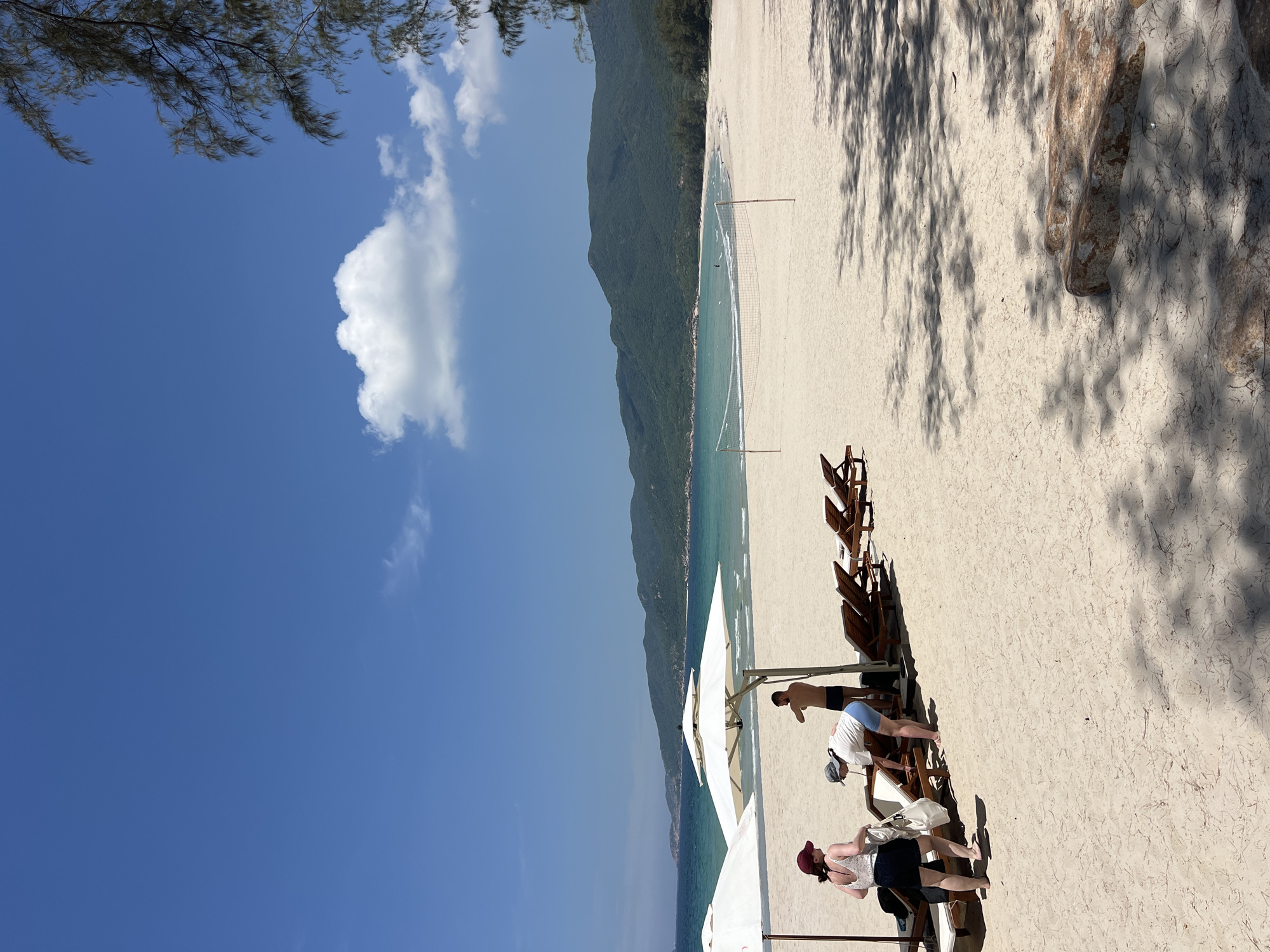 A sunny, clear day at a beach resort in Cong Hai, Vietnam. The sandy shore is lined with empty lounge chairs and white umbrellas. In the foreground, three people are visible near the umbrellas. One person, wearing a white top and blue shorts, is bending over a lounge chair. Another person, with a red cap and white top with black trim, is standing and holding a white bag. A third person, a shirtless male in black swim trunks, stands between them. Further down the beach, a volleyball net is set up on the sand. The turquoise ocean stretches out to a distant, green, mountainous coastline. The sky is a vibrant blue with a few scattered white clouds. Tree branches cast dappled shadows on the sand in the lower right corner of the frame.