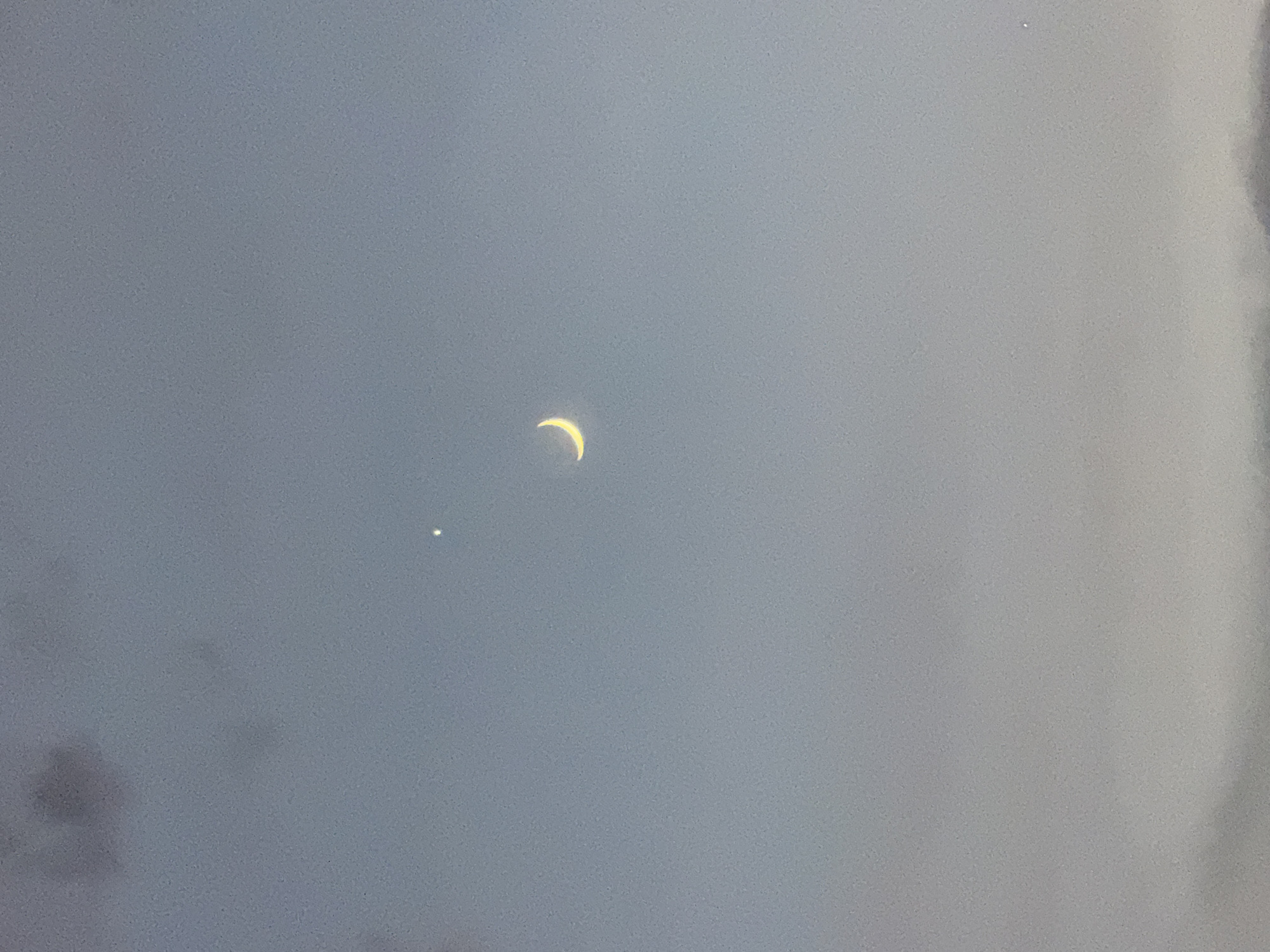 In the twilight sky above Rafailovići, Montenegro, a sliver of the crescent moon hangs prominently, its pale yellow glow contrasting with the muted blue of the fading day. A bright point of light, likely a planet, is visible higher in the sky. The lower portion of the image is obscured by soft, hazy clouds or mist, obscuring any view of the town or the Adriatic Sea. The overall impression is one of celestial solitude and the quiet transition from day to night.