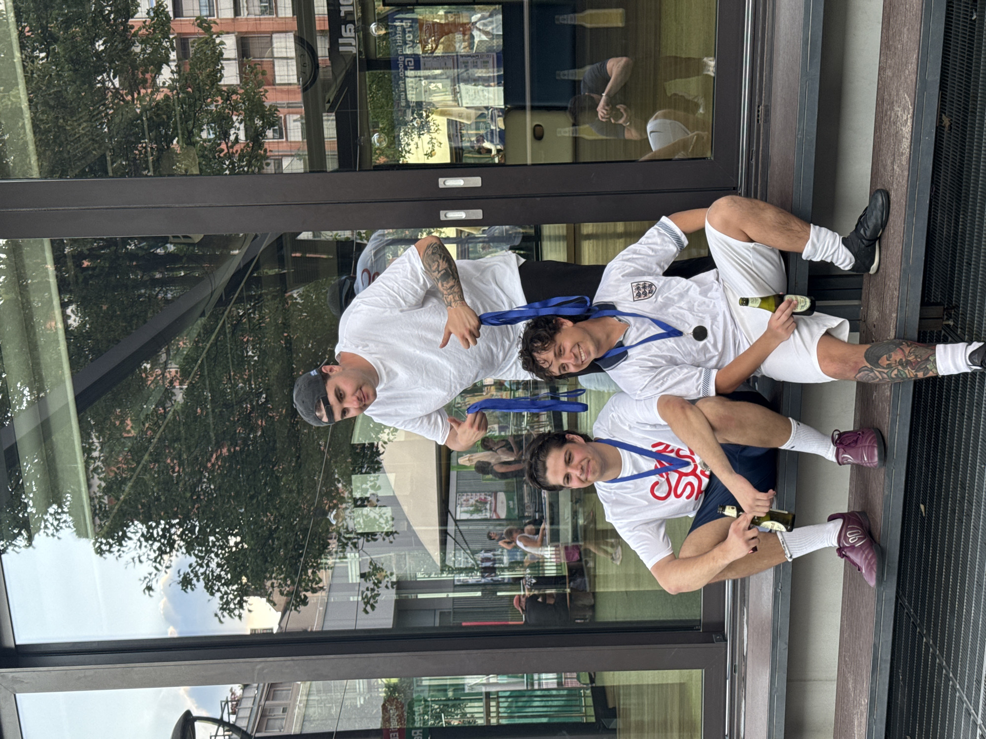 The image depicts three adult males positioned on dark grey metal steps in front of a modern building with a dark-framed, highly reflective glass facade, located in an urban setting in Milan, Italy.
The central figure, standing behind the other two, has dark hair, wears a dark baseball cap backward, a white short-sleeved t-shirt, and dark trousers. Visible tattoos adorn his left forearm. He smiles and extends both arms forward, giving a thumbs-up with both hands while holding blue straps or ribbons.
To the left, seated on the steps, is a male with dark curly hair. He wears a white short-sleeved t-shirt with red lettering on the chest, partially visible as "SUN SOAKED." He also wears dark blue shorts, white crew socks, and dark purple athletic shoes featuring the numeral "90" on the lateral side of the left shoe. A blue ribbon with a silver-colored medal is suspended around his neck. He smiles and holds a green glass beer bottle in his right hand.
To the right, also seated on the steps, is another male with dark curly hair. He wears a white short-sleeved England national football team jersey, displaying the Three Lions crest. He is dressed in white shorts, white crew socks, and black athletic shoes. His left calf exhibits extensive tattooing. A blue ribbon with a gold-colored medal is around his neck. He smiles and holds a green glass beer bottle in his left hand.
The reflective glass facade behind the individuals displays various elements of the surrounding environment. In the upper left, a bright sky with distinct clouds is reflected. Extensive green tree foliage is prominent across the upper and middle sections of the reflection. The upper right reflection shows a multi-story brick-red building with numerous windows. Other urban structures and partial, inverted signage, including "JJS 10" and "ELIB," are also visible. Reflections of several other individuals are present within or beyond the glass, including one person in a red cap seated at a table on the left, and a person in the lower right appearing to hold a device. The outlines of two beer bottles standing on a shelf inside the building are reflected in the lower right.