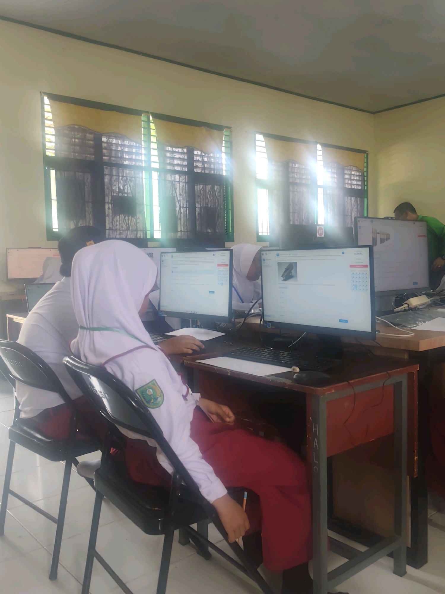 In what appears to be a classroom in Pematang Reba, Indonesia, several students are seated at desks facing computer monitors. The room is brightly lit by sunlight streaming through windows with yellow curtains. Two students in the foreground are wearing uniforms: one in a white shirt and maroon skirt, and another in a white hijab and white shirt with a maroon skirt. Both are looking at their computer screens. One student's head is bowed, and the other is actively engaged with the computer. A third student, partially visible, is also at a computer. In the background, a teacher or administrator in a green shirt is standing and looking at a monitor. The desks are simple wooden structures, and the floor appears to be tiled. The overall impression is one of a school computer lab during a lesson or exam.