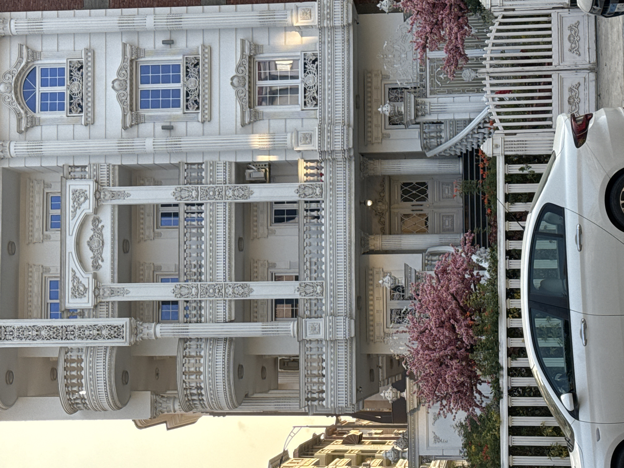 A photograph captures a meticulously ornamented white facade of a building, potentially a house, set against a blurred cityscape in the background. The architecture exhibits intricate detailing, including arched windows, elaborate columns, and decorative balustrades, conveying an impression of luxury and attention to detail. A blooming tree with pink blossoms is positioned in the foreground, adding color and life to the scene. A white car is parked beside a white picket fence, adding another layer to the composition. The photograph's focus on the architectural details and the car beside the home, suggests a residential setting.