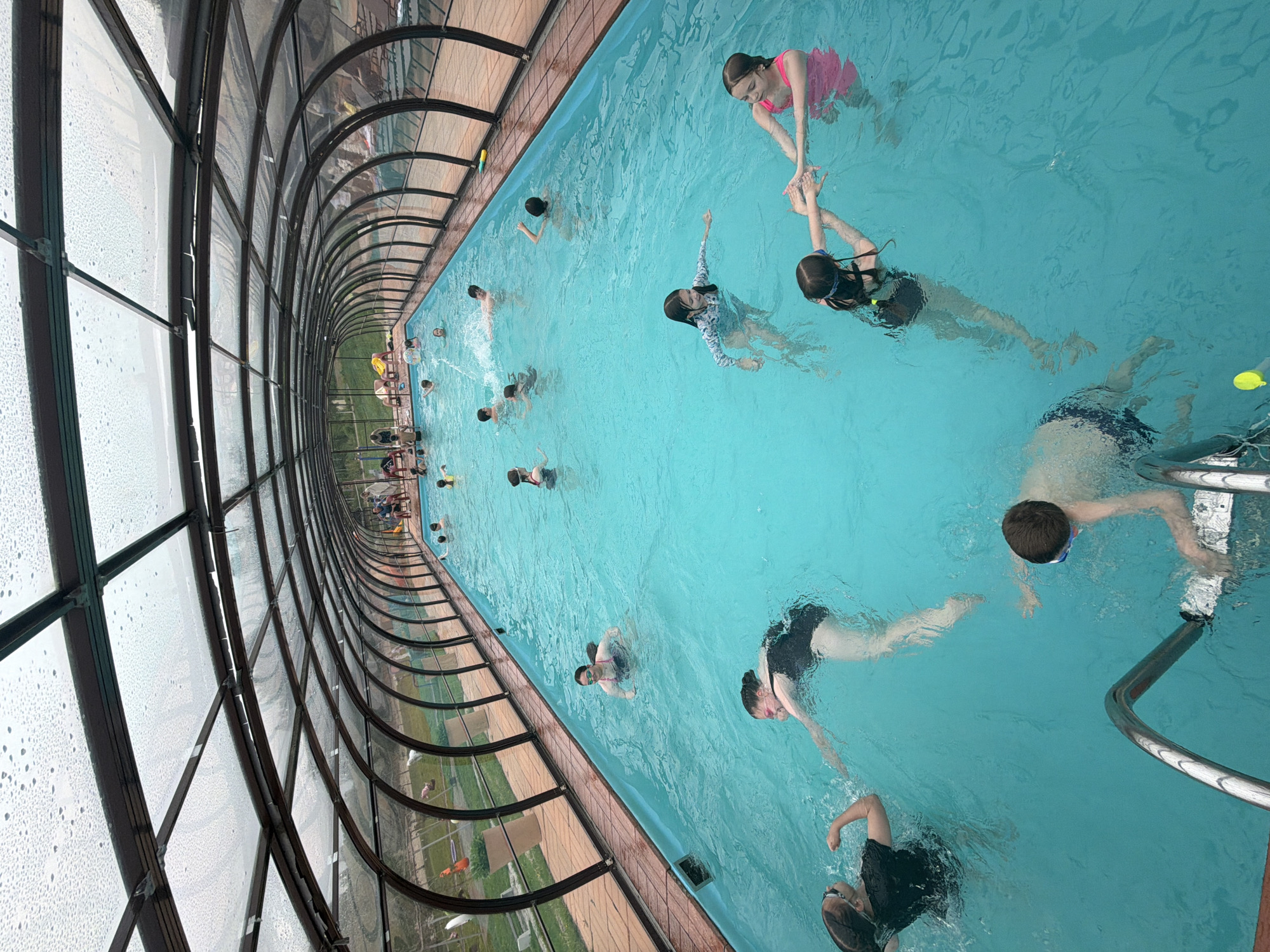 A high-angle shot depicts a rectangular indoor swimming pool filled with people, situated in what appears to be a covered or enclosed area, possibly in the city of Birobidzhan, Russia. The pool is predominantly light blue and contains at least a dozen individuals of varying ages. Several people are actively swimming, while others seem to be standing or floating in the water. In the upper right corner, two young girls can be seen holding hands. One is submerged, and the other is supporting the swimmer.
The pool is surrounded by a distinctive arched glass and metal structure. This structure allows natural light to enter, but the glass appears to be wet with condensation. Beyond the structure, glimpses of green grass and trees are visible, suggesting an outdoor environment adjacent to the pool area. A metal ladder is present in the pool's corner. The scene appears to capture a recreational swimming activity in a controlled environment.