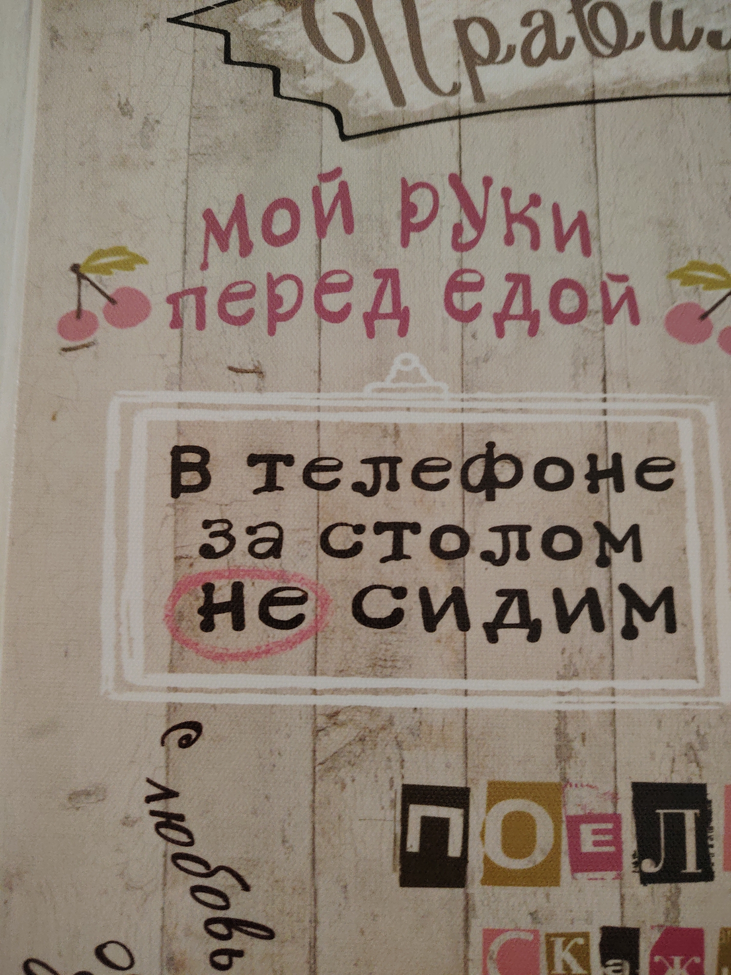 The image displays text in Russian on a light-colored, textured background that resembles wood paneling. In the upper portion of the image, a large, ornate script reads "Правила" (Rules). Below this, in a pink, stylized font, are the phrases "мой руки" (my hands) and "перед едой" (before eating).
Centered in the image is a white rectangular frame containing further text in a black, blocky font. This text reads: "В телефоне" (In the phone), "за столом" (at the table), and "не сидим" (don't sit). The word "не" (not) is circled in pink.
To the left of the framed text, a handwritten phrase in cursive script reads "с любовью" (with love). In the lower portion of the image, a series of colored square blocks with letters are visible, spelling out parts of words. The visible letters form "ПОЕЛ" (ate) and fragments of other words.
The image does not contain any information that would allow for its location in Vladimirovka, Russia, or any other specific city. There are no people, actions, or interactions depicted. The scene is a static display of text and graphic elements.