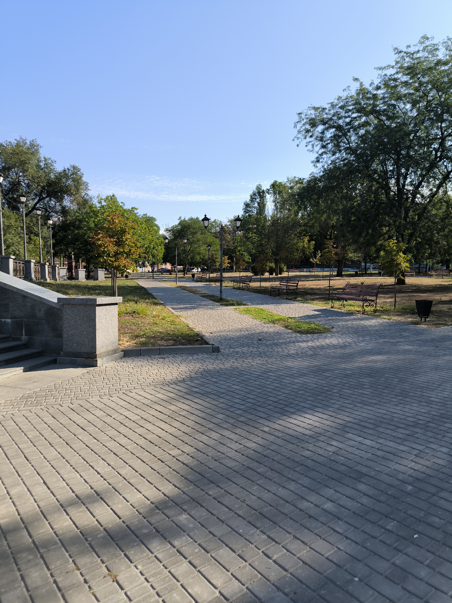This is a photograph of a public park in Mykolaiv, Ukraine. The image shows a paved walkway leading into the park, lined with trees and benches. The sky is a clear blue. There are no people visible in the image.
