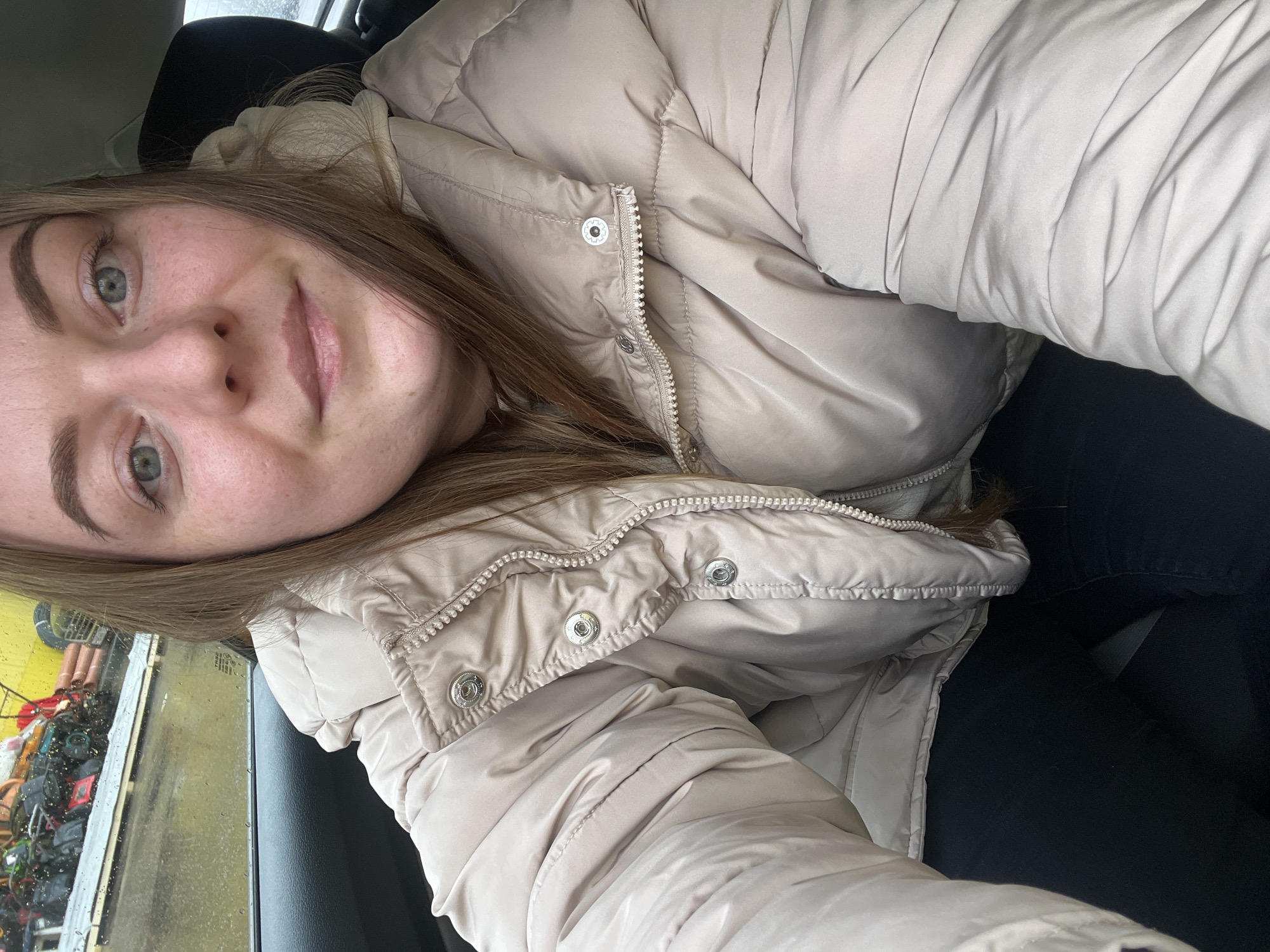 A young woman is taking a selfie inside a vehicle. She has fair skin, light brown hair, and is wearing a beige puffy winter coat. Rain is visible on the car window, and through the window, various objects are stacked in what appears to be an outdoor storage or work area. No specific details indicate the location is Shevlyakovo, Russia.