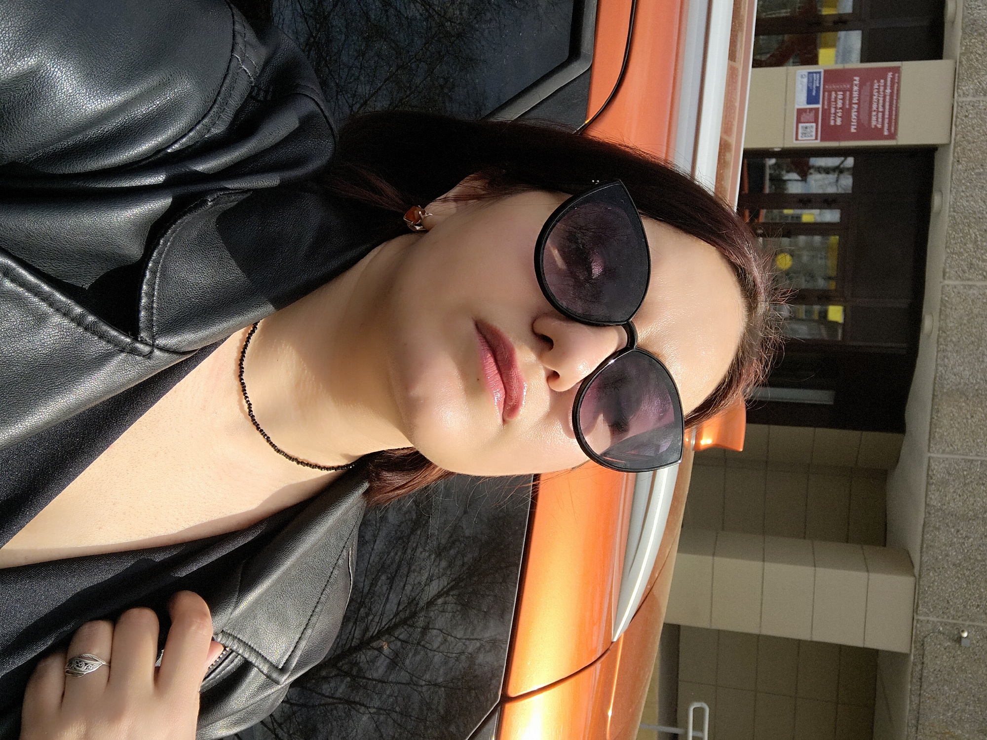 A woman in an unzipped black leather jacket stands next to an orange car in Марковский, Russia. She wears black sunglasses and a black beaded necklace. Her hand with a ring is near the zipper. A building with brown doors and signs is visible in the background.