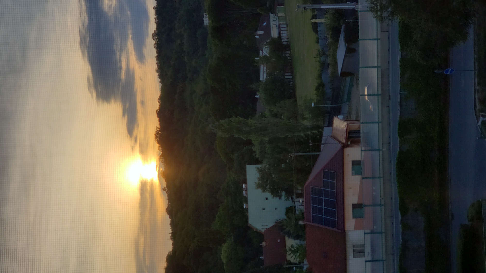 The image depicts a serene sunset scene over a semi-urban landscape. The sun, a bright orb radiating golden light, is partially obscured by clouds and the crest of a densely wooded hill. The sky transitions from a soft yellow near the horizon to a pale blue and grey as it ascends. The hill, covered in lush green trees, dominates the background, with glimpses of houses nestled within its slopes. In the foreground, several buildings are visible. One prominent structure features a red roof with an array of solar panels. Adjacent to it, another building is painted in lighter colors and has visible windows. A clear fence separates the buildings from a road, on which a silhouetted car is visible. The overall atmosphere is peaceful and picturesque, capturing the calm beauty of a suburban evening.