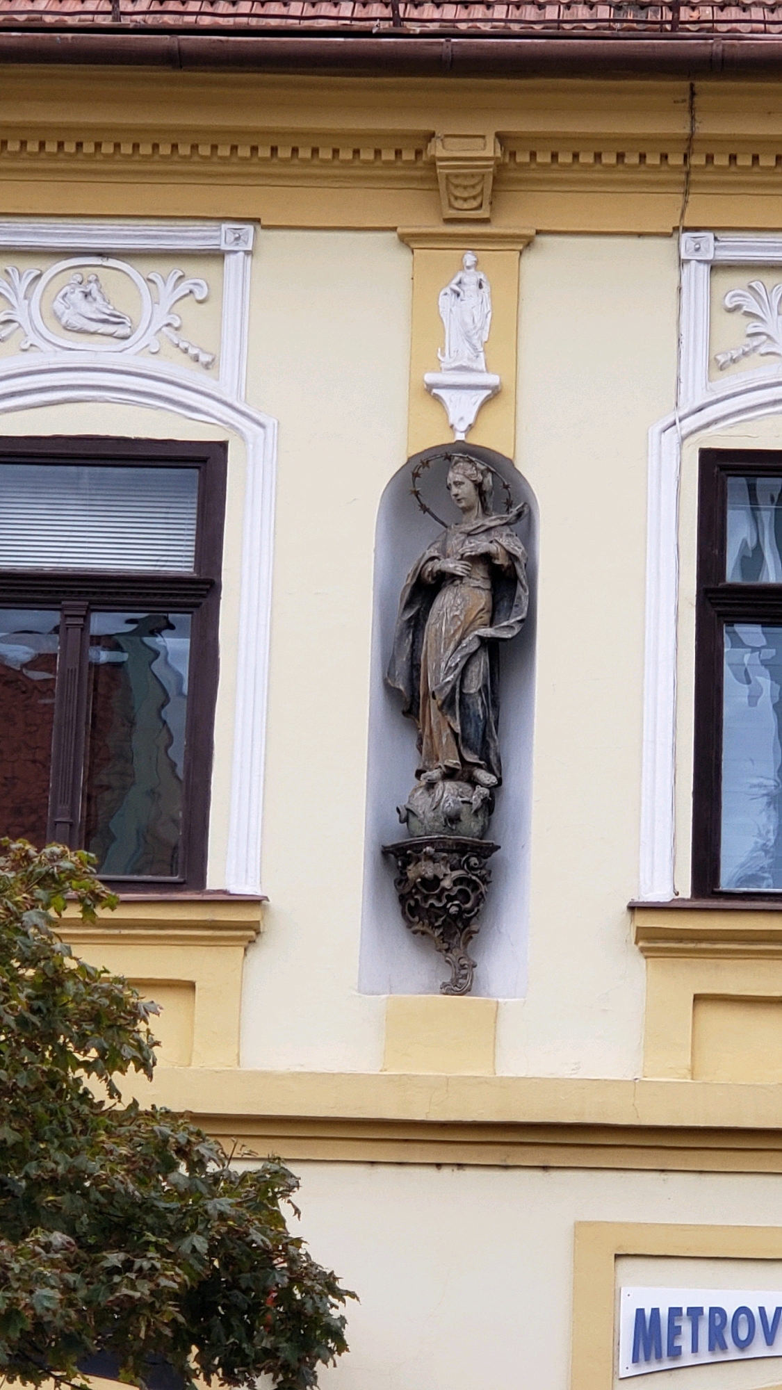 This is a low-angle medium shot of the facade of a building in Prešov, Slovakia, featuring architectural details and a statue. The building has a light yellow facade with darker yellow decorative trim. To the left, a dark brown window with white blinds is visible, framed by white decorative molding. To the right of the window, a recessed alcove contains a full-body statue of a woman with a halo of stars. She is wearing a draped robe and holding an object in her hands. The statue stands on a globe, which rests on an ornate dark metal bracket. Above the alcove, a smaller white statue of a figure standing on a pedestal is visible. Further to the left and right of the central alcove, there are other decorative elements, including a white relief sculpture above the window on the left. The top of the building is adorned with a brown tiled roof. A section of a green leafy tree is visible in the lower left corner of the frame. In the lower right corner, a white sign with blue lettering reads "METROV".