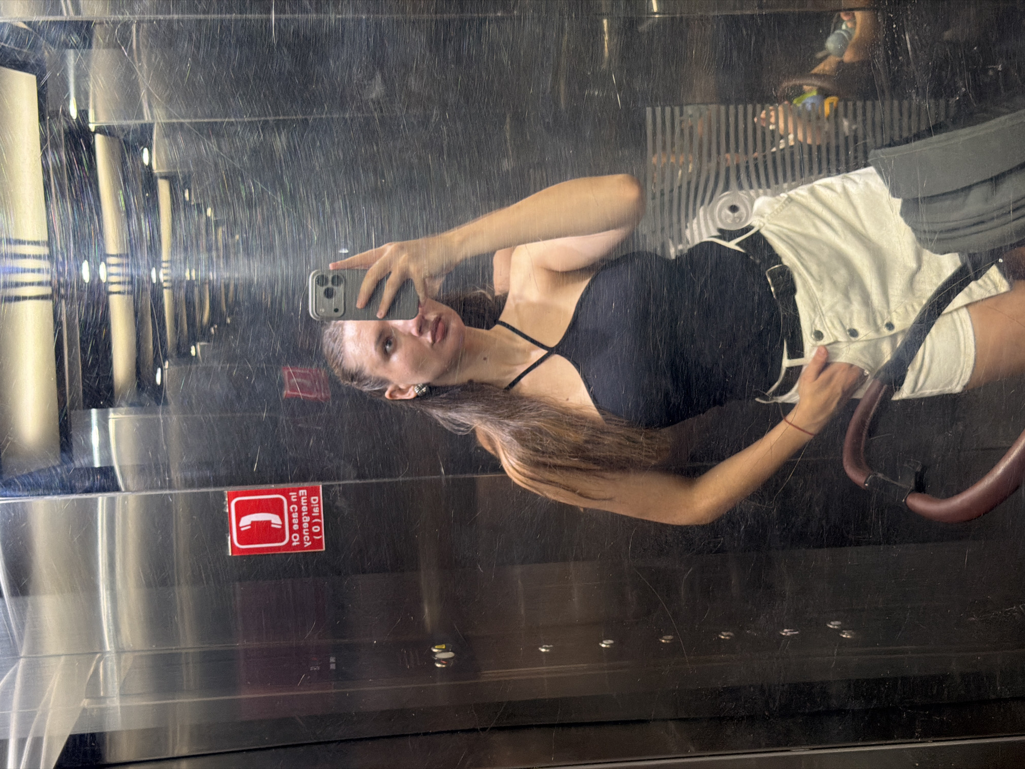 A young woman with long, dark brown hair pulled back into a ponytail, wearing a black halter top and white shorts, takes a mirror selfie in an elevator. She is holding a gray smartphone in her right hand. The elevator walls are made of reflective stainless steel, showing streaks and scratches. On the left wall, a red emergency sticker with a phone icon and text in a foreign language is visible. In the lower part of the image, a stroller handle with a maroon grip is partially visible. The lighting in the elevator appears to be from overhead fluorescent fixtures. The scene is indoors, within what appears to be a standard elevator. The city of Sharm El Sheikh, Egypt, is a tourist destination known for its beaches and diving sites, but this specific image does not contain any visual elements that directly indicate this location.