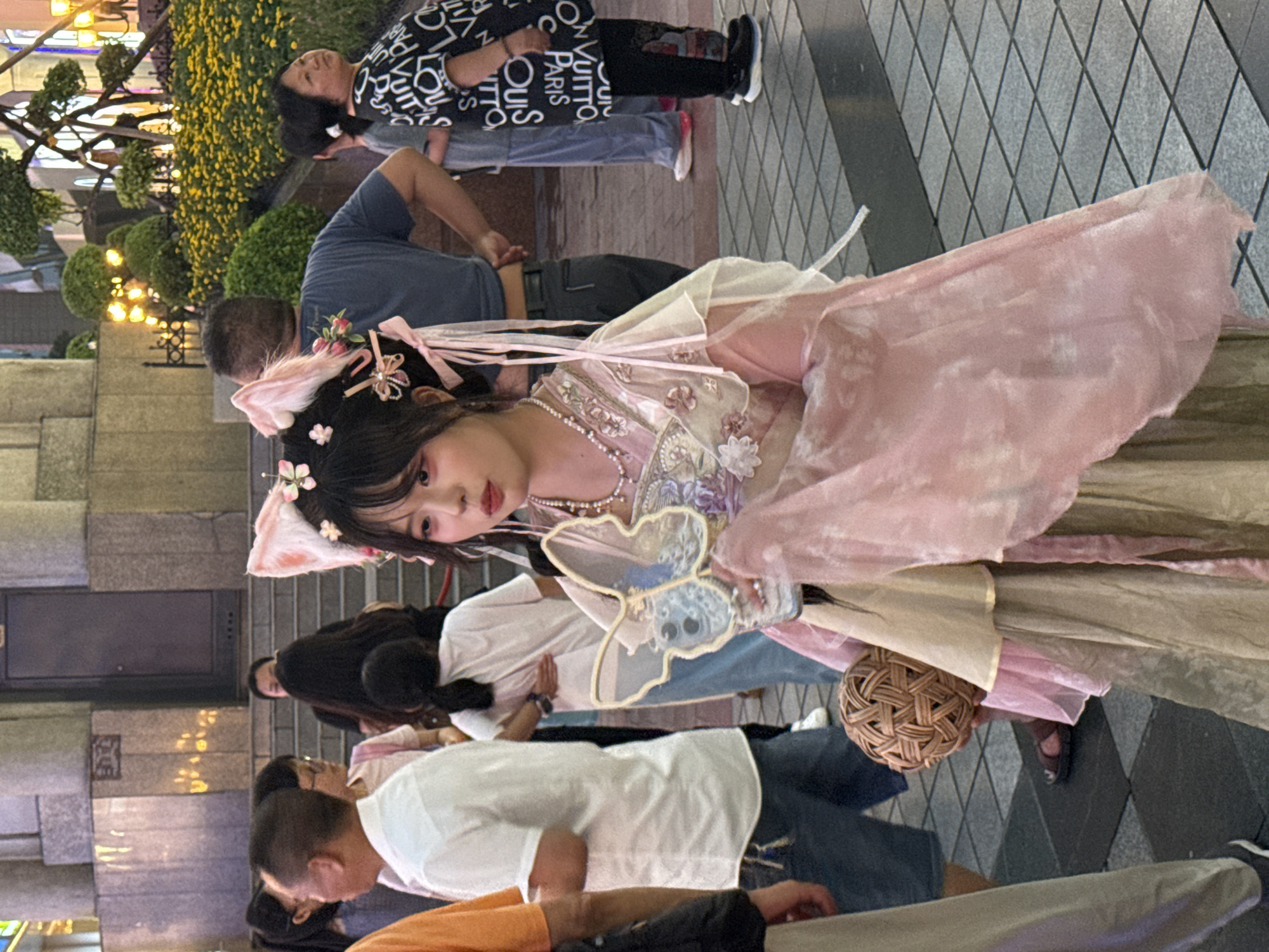The image shows a young woman dressed in a pink and gold traditional Chinese garment, possibly a Hanfu, with cat ears headband. She is holding a butterfly-shaped fan and a woven ball. She is standing on a city street in Yuzhong District, China, at night. The background shows other people, streetlights, and buildings. The woman is the main subject of the image. She is wearing heavy eye makeup and her hair is styled with ribbons and flowers. Her outfit appears to be for a costume or performance. The surrounding environment suggests a busy urban area with a mix of modern and traditional architecture. The lighting is a mix of artificial streetlights and ambient city glow.