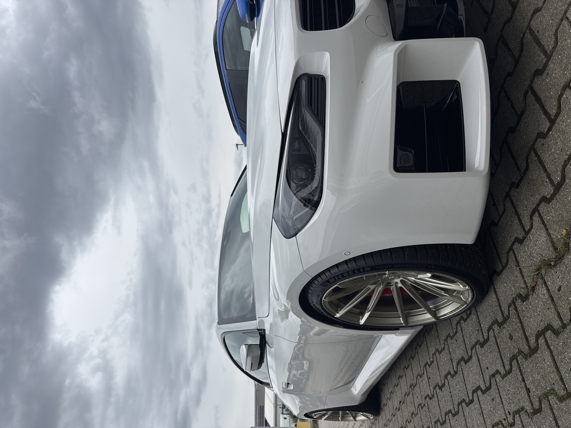 The image shows the front passenger side of a white BMW M2 CS. The car is parked on a grey, interlocking brick driveway. The sky is overcast, suggesting a cloudy or rainy day. In the background, a blue car is visible, also parked. The BMW has a large, aggressive front bumper with prominent air intakes. The headlight is visible, featuring a modern LED design. The car is equipped with a gold, multi-spoke wheel and a Michelin Pilot Sport 4S tire. The brake caliper is red. The BMW logo is visible on the wheel. The scene appears to be a car parking area, possibly in Wettstetten, Germany, given the location context. There is no discernible activity taking place, as the cars are stationary. The overall mood is neutral, with the focus on the car's design and details.