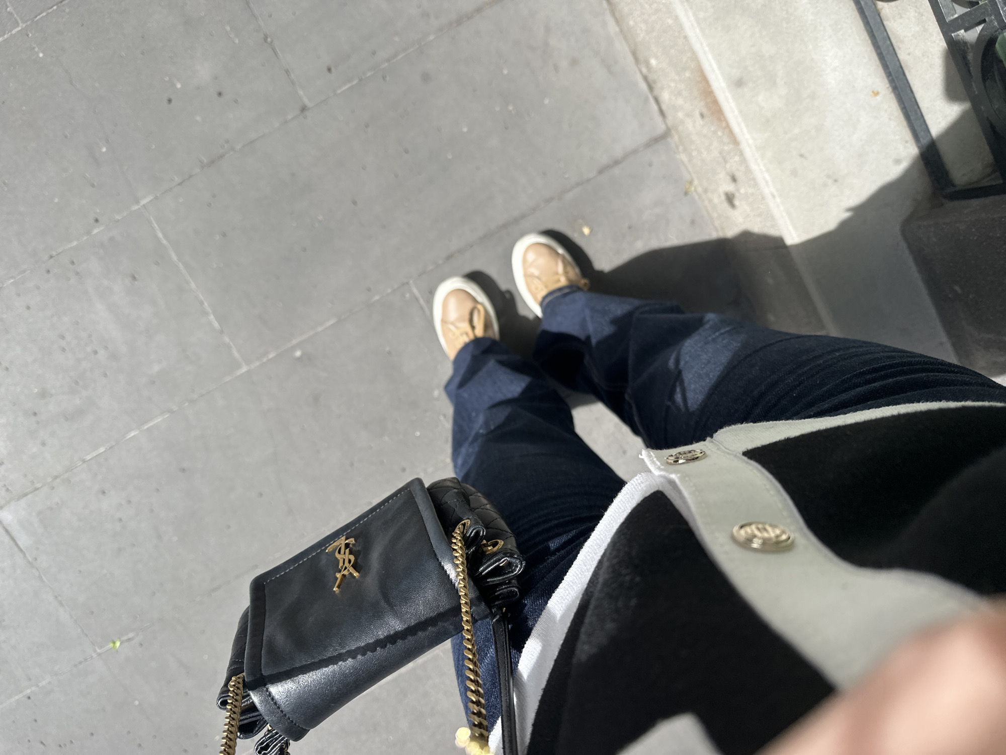 A top-down view captures a person standing on a paved surface, likely a sidewalk, with a portion of a building's base visible in the upper right corner. The person is wearing dark blue jeans and light tan shoes. A small black leather purse with a gold chain strap and a gold emblem is held in their left hand. The person is also wearing a black and white cardigan, with the right side of the garment prominently displayed. The scene is illuminated by sunlight, casting shadows on the pavement. Based on the architectural details and the style of paving, the location could potentially be in Yerevan, Armenia.