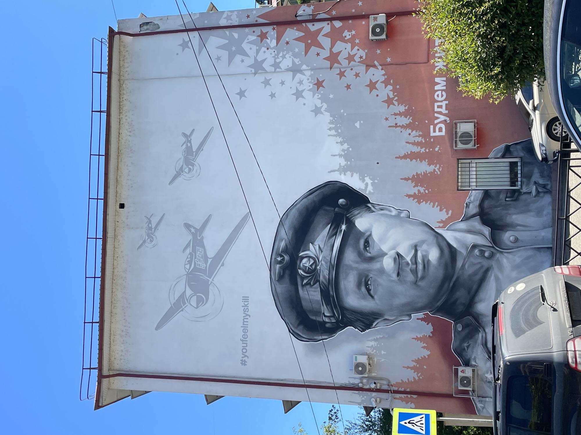 A large outdoor mural dominates the side of a building in Sochi, Russia, depicted under a clear, bright blue sky. The central feature of the mural is a large, black-and-white portrait of a young man, likely a pilot or military figure, wearing a cap with a star emblem. His expression is serious, and his gaze is directed forward.
Behind and above the man, the mural transitions to a background composed of stylized pine tree silhouettes in reddish-brown, which blend into a pattern of stars in both reddish-brown and gray tones. To the left of the portrait, several gray, propeller-driven fighter planes are depicted in various flight positions.
Visible text on the mural includes the English hashtag "#youfeelmyskill" positioned below the planes, and the Russian phrase "Будем жить!" (translating to "We will live!") written vertically in white on the reddish-brown section of the wall.
The building's light-colored facade shows some weathering and dirt. Several white air conditioning units are mounted on the wall, integrated into the mural's design. A tall, thin metal ladder or railing runs along the left edge of the building.
The lower right portion of the image is partially obscured by real-world elements, including parts of two parked vehicles: a silver SUV (with "LAND CRUISER" and "V6" visible on its rear) and a darker-colored van. A green leafy bush or tree and a blue and white pedestrian crossing sign are also visible in the foreground. The scene is brightly lit, indicating daytime conditions.