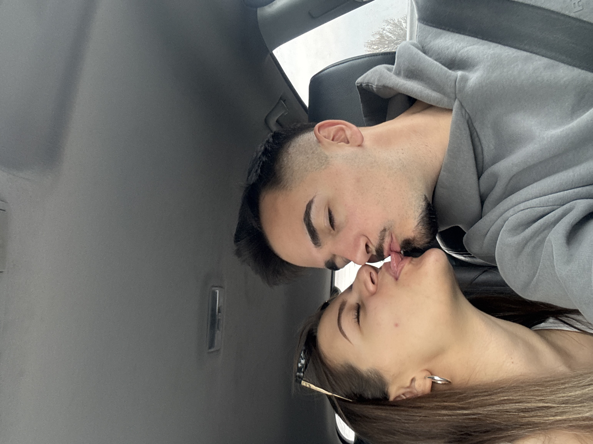 A photograph depicts two individuals, a male and a female, kissing in what appears to be the interior of a car. The interior is gray. The male is wearing a gray hooded sweatshirt. The image is oriented in a portrait format but appears to be rotated clockwise, suggesting the subjects are reclined. The location, Cherna Gora, Bulgaria, is not discernible from the visual information available.