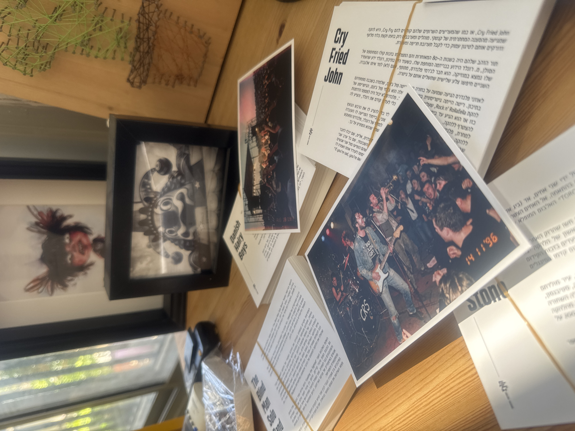 This image captures a static display on a wooden surface, likely a desk or table, under natural light during the daytime. The arrangement features several paper booklets or pages, two framed pictures, and a piece of string art in the background.
The central focus is on a collection of printed materials, predominantly in Hebrew, which appear to be related to music, possibly fanzines, a book, or promotional material. One booklet is clearly titled "Cry Fried John" in English, with accompanying Hebrew text discussing a band from the "underground scene of Kentucky" that plays "kicking rock and a bit of caressing blues." Another piece of paper mentions "Danish Hairy Boys," and "The Blind Man Solo Band" is also visible. Several photos embedded in or alongside the text depict live music performances: one shows a band on stage with a guitarist wearing a denim jacket with "BAD LIDA GIFT" and "CRS" printed on it, and a date "14 11 96" is visible in the background. Another photo shows a concert crowd. An older photo, partially obscured, shows a date "AUG 18 1969". Extensive Hebrew text fills the pages, discussing band histories, musicians like "Ronald," and musical styles. "Stone" and "TORC" are also partially visible near the bottom right.
To the left, two framed pictures are visible. The top one, partially obscured, is a black and white artwork featuring an abstract or cartoonish face. Below it, another framed picture shows a blurred, abstract red and black pattern, possibly another artwork or a cropped image.
In the background, a light wooden surface holds a string art piece with green and dark-colored strings forming geometric patterns. A reflection from a window is faintly visible on the left side, indicating an indoor setting with natural light. The presence of Hebrew text strongly suggests a location in Israel, consistent with the provided context of Tel-Aviv. The overall impression is a personal collection or a display with a strong focus on music and its history.