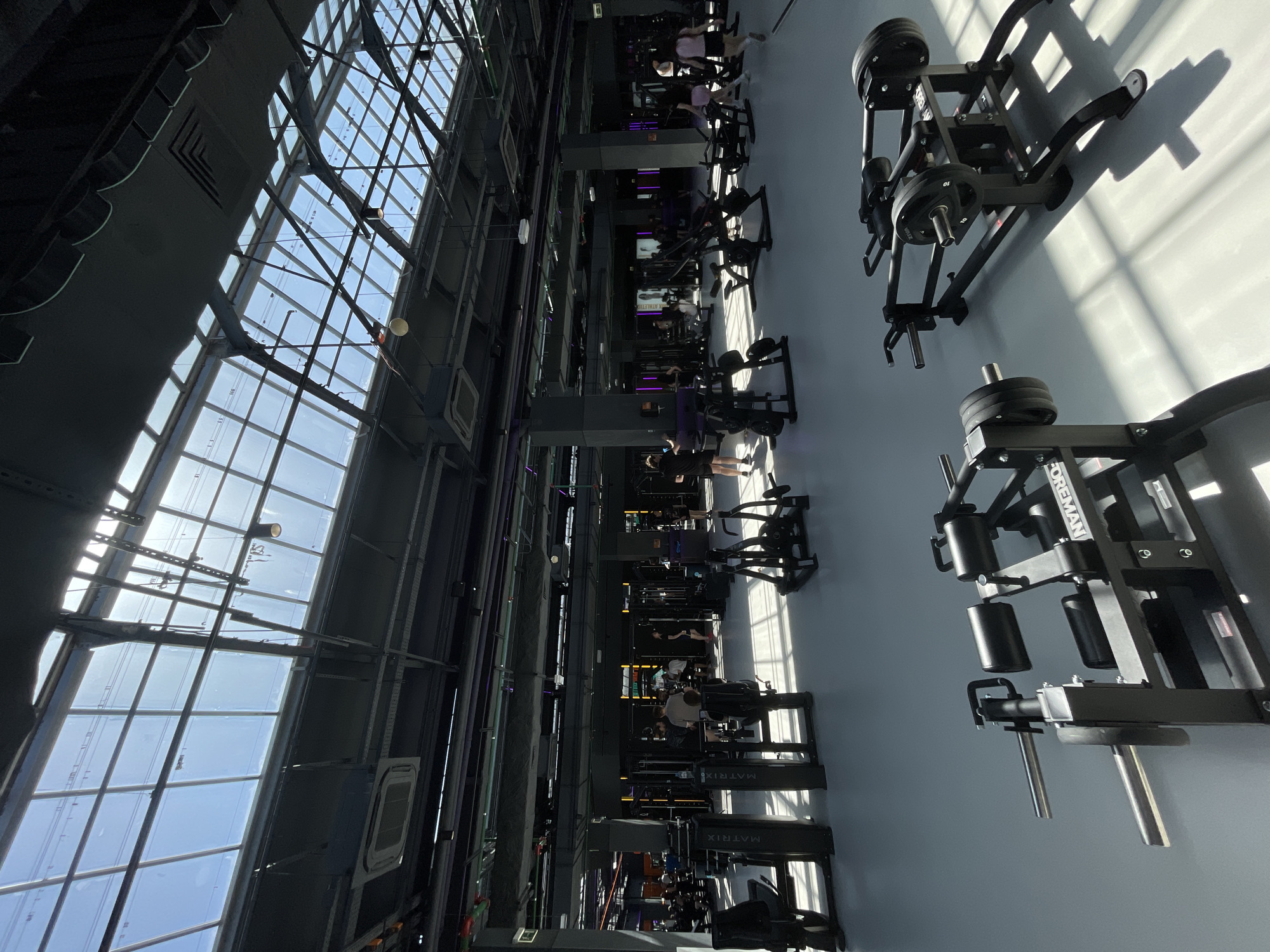 This image shows the interior of a gym with a high ceiling and a large window wall that lets in natural light. The equipment is lined up in rows, with people using the machines. The brand Matrix is visible on the equipment. Based on location, this is likely Planet Fitness in Kazan, Russia.
