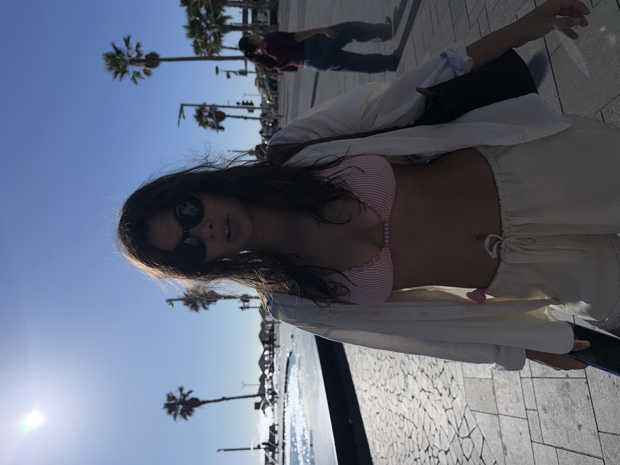 A young woman with long, dark hair, wearing sunglasses, a pink and white striped bikini top, and a light-colored, open button-down shirt, poses for a photo outdoors. She is wearing loose-fitting, cream-colored shorts. The image is taken from a low angle, with the sky and palm trees in the background. The sun is bright, suggesting it is daytime, likely late morning or afternoon. She appears relaxed and is looking at the camera.
In the background, a person in a dark shirt and jeans is walking away. There are more palm trees and what appears to be a promenade or walkway with a patterned stone surface. Further in the distance, there are buildings and a street with traffic lights. The scene suggests a warm, sunny day in a coastal or resort-like setting. The presence of palm trees and the light are consistent with a location like Tel Aviv, Israel. The water feature in the foreground, with its reflections and shimmering surface, adds to the summery feel. The overall mood is casual and cheerful. There is no visible text in the image.