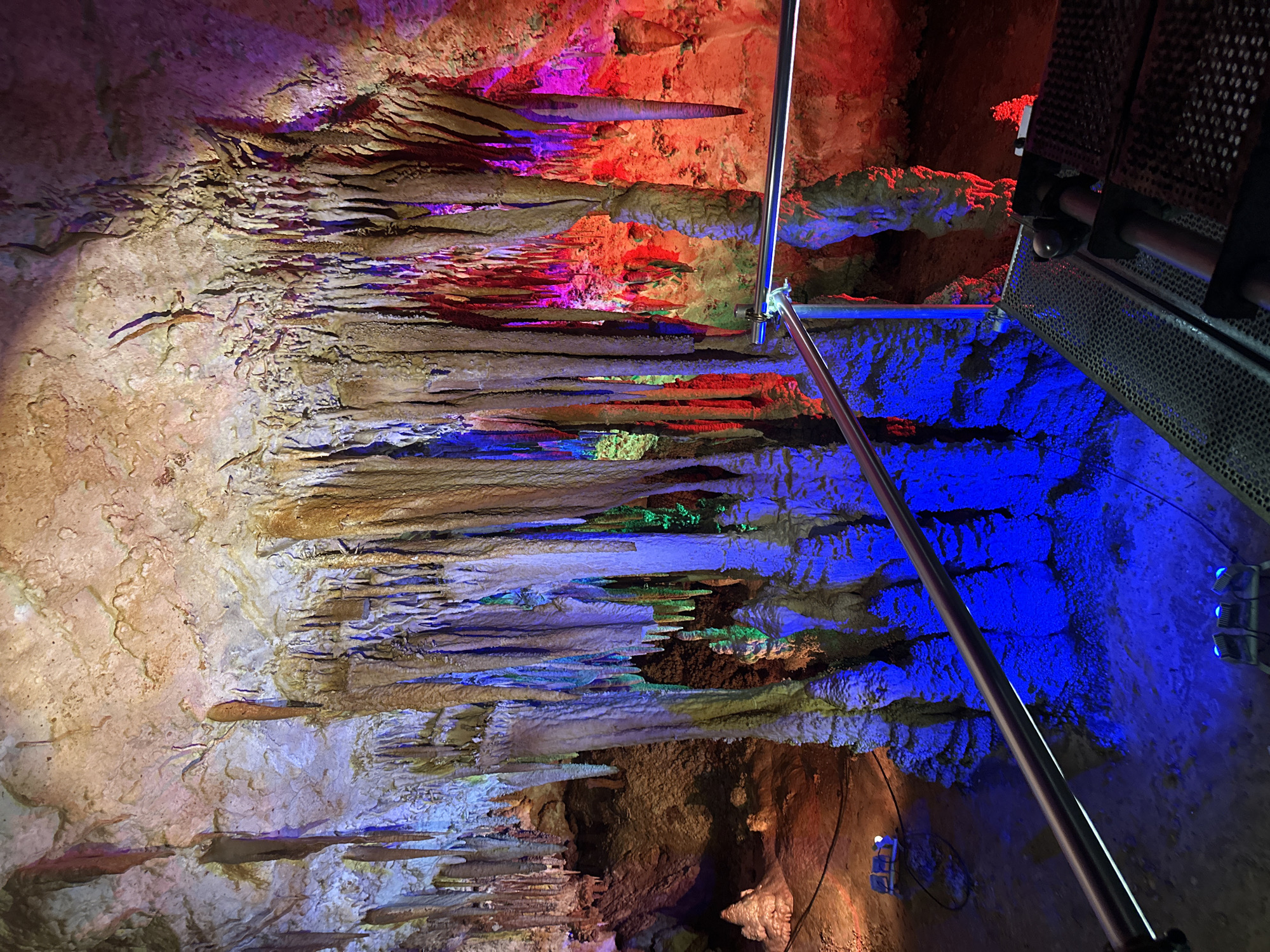 The image shows the interior of the Uhlovitsa cave in Belo pole, Bulgaria. The cave walls and ceiling are covered with stalactites and stalagmites, illuminated with colored lights. The colors include red, purple, green, and blue. A metal railing and metal grated pathway are visible on the right side of the image.