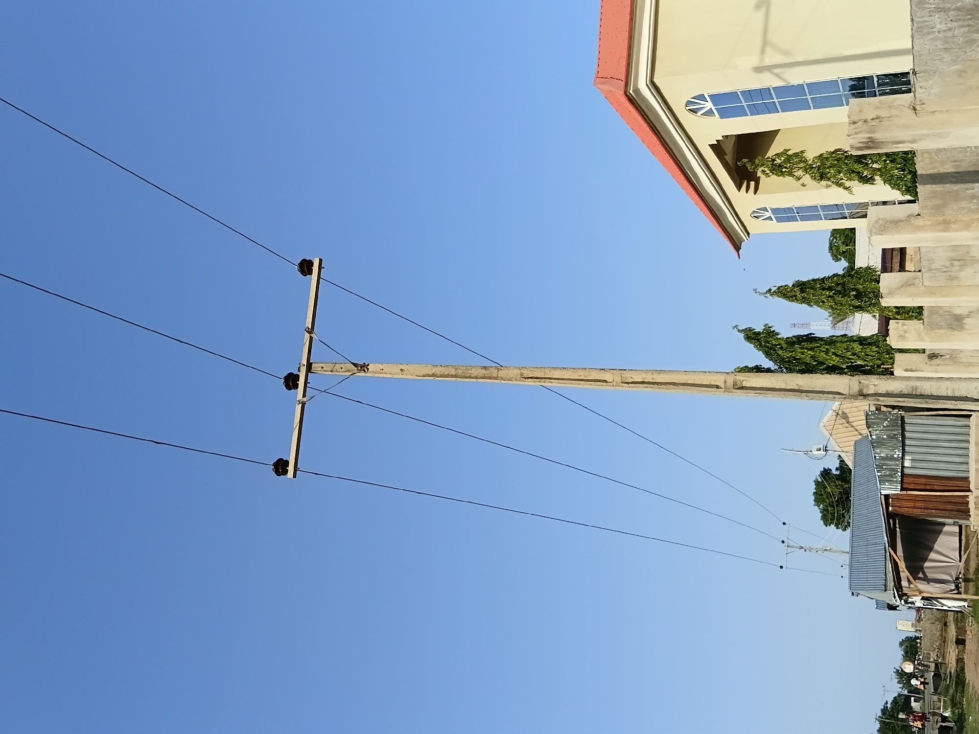 In Jalingo, Nigeria, a concrete utility pole stands tall against a clear blue sky, supporting multiple electrical wires that stretch across the frame. To the right, a light-yellow building with a red roof and arched windows is partially visible, bordered by a low concrete wall and several tall, slender trees. In the middle ground, several corrugated metal structures with varying rooflines and wooden supports are clustered together, suggesting a commercial or residential area. Further to the left, a paved road runs alongside the structures, with a few indistinct figures walking on the sidewalk. The scene is illuminated by natural daylight, casting subtle shadows and highlighting the textures of the materials present.