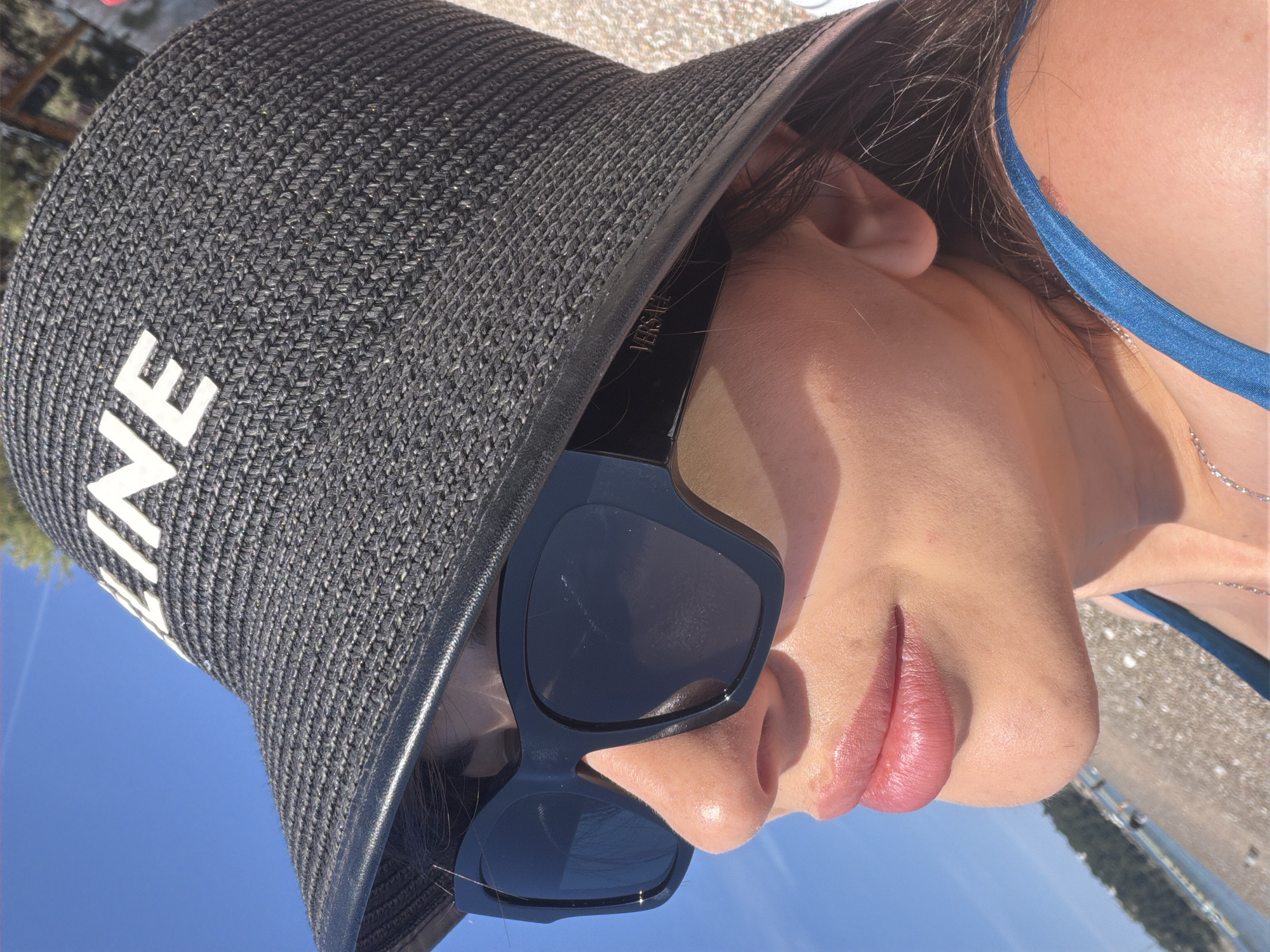 The image is a close-up, rotated portrait of a person, likely a woman, seen from the nose up at an upward angle. She is wearing a black woven sun hat with a wide brim, featuring the white capital letters "ELINE" (part of "CELINE") on its side. Large, rectangular black sunglasses cover her eyes, with the word "VERSACE" visibly inscribed in gold on the right temple. Her lips are full and glossy pink. Dark hair is visible around her ear and under the hat. A blue strap, appearing to be from a bikini top, is visible on her right shoulder area, alongside a delicate silver chain necklace.
The background shows a clear, bright blue sky on the upper right side. The lower left of the image (when rotated) reveals a sunlit pebbled or rocky beach, leading to calm water and distant green landmasses, indicating a coastal setting. The strong, direct sunlight suggests a sunny day, likely mid-day, creating bright highlights on her skin and reflections on the sunglasses. The overall scene conveys relaxation in a warm, outdoor environment. The location is Rafailovići, Montenegro.