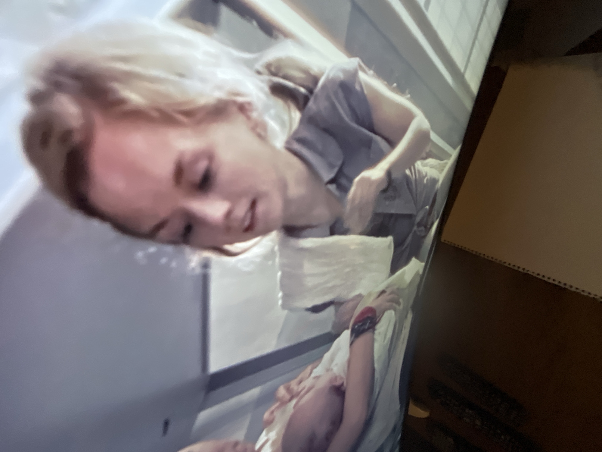 A fair-skinned woman with blonde hair, wearing a grey top, is looking down at a baby wrapped in a white blanket in what appears to be a medical setting. The scene may be from a television screen, given the angle and surrounding elements. The surrounding environment includes medical equipment or a hospital bed and a wooden table or wall. There is no indication that the location is San Bernardino Tlaxcalancingo, Mexico.