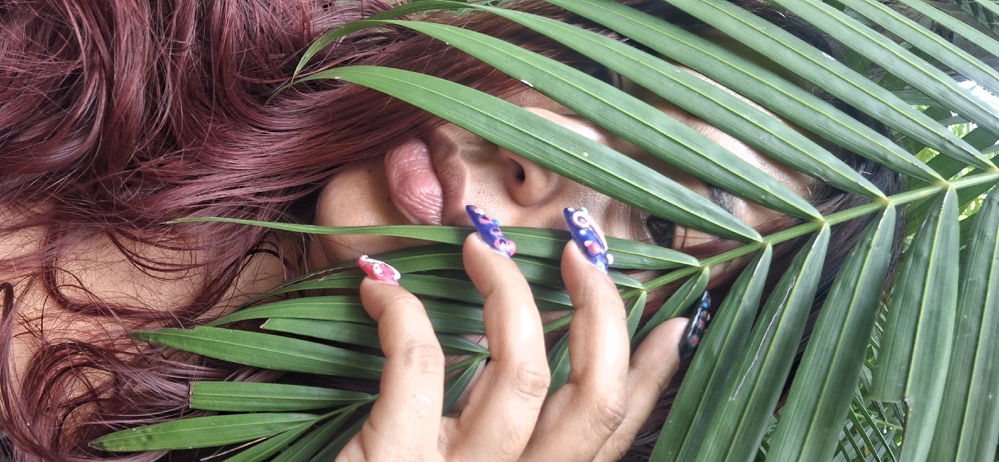 A close-up, horizontal shot depicts a person partially obscured by large, vibrant green palm-like leaves, set in what appears to be an outdoor, natural environment consistent with Vallecito, Venezuela.
The subject's face is mostly hidden by the foliage, with prominent reddish-burgundy, wavy hair visible on the left side of the frame. Through the leaves, parts of the person's nose and one eye are discernible. Playfully, a tongue is extended from their mouth, adding a lighthearted or mischievous element to the expression.
Fingers are visible, holding some of the leaves, and drawing attention to their elaborately decorated nails. The nails are long, pointed, and feature distinct, colorful designs: one finger shows a pink nail with white details, while others display purple and black nails adorned with vibrant dots and abstract patterns.
The image emphasizes a contrast between the organic green of the leaves and the rich, artificial color of the hair and the playful artistry of the nails. The natural lighting suggests it is daytime. No text is visible in the media.