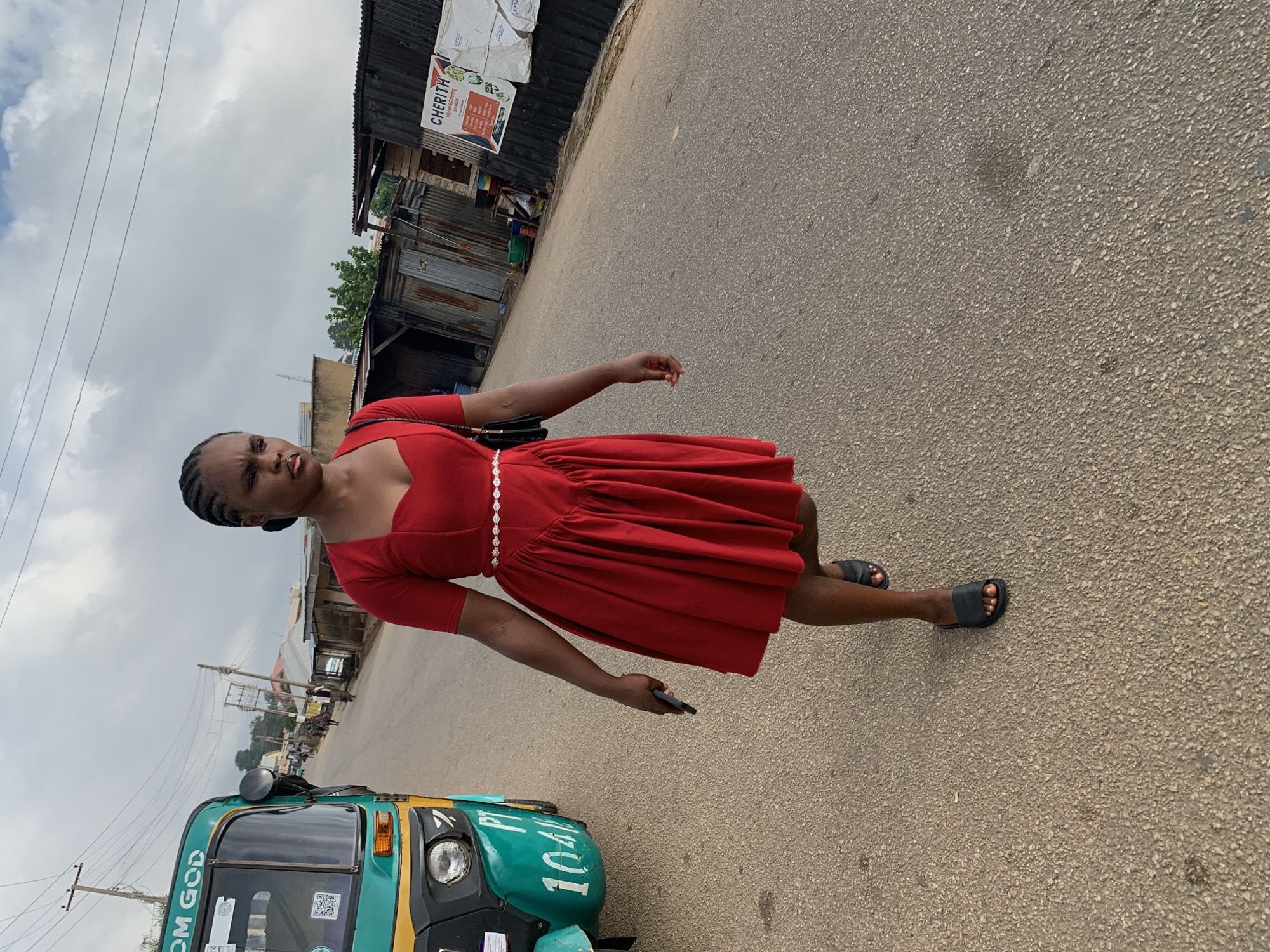 A young woman with braided hair, wearing a red off-the-shoulder dress with a sweetheart neckline and a full skirt, walks on a paved road in Jos, Nigeria. Her left arm is bent, and she holds a dark-colored mobile phone in her left hand. She wears black slide sandals. To her left, a green and yellow auto-rickshaw, identified with "OM GOD" and "PT 1041" on its front, is parked. The background shows a street with various structures, including corrugated metal shops and a billboard with the text "CHERITH Kitchen & Catering Service". The sky is overcast with clouds.