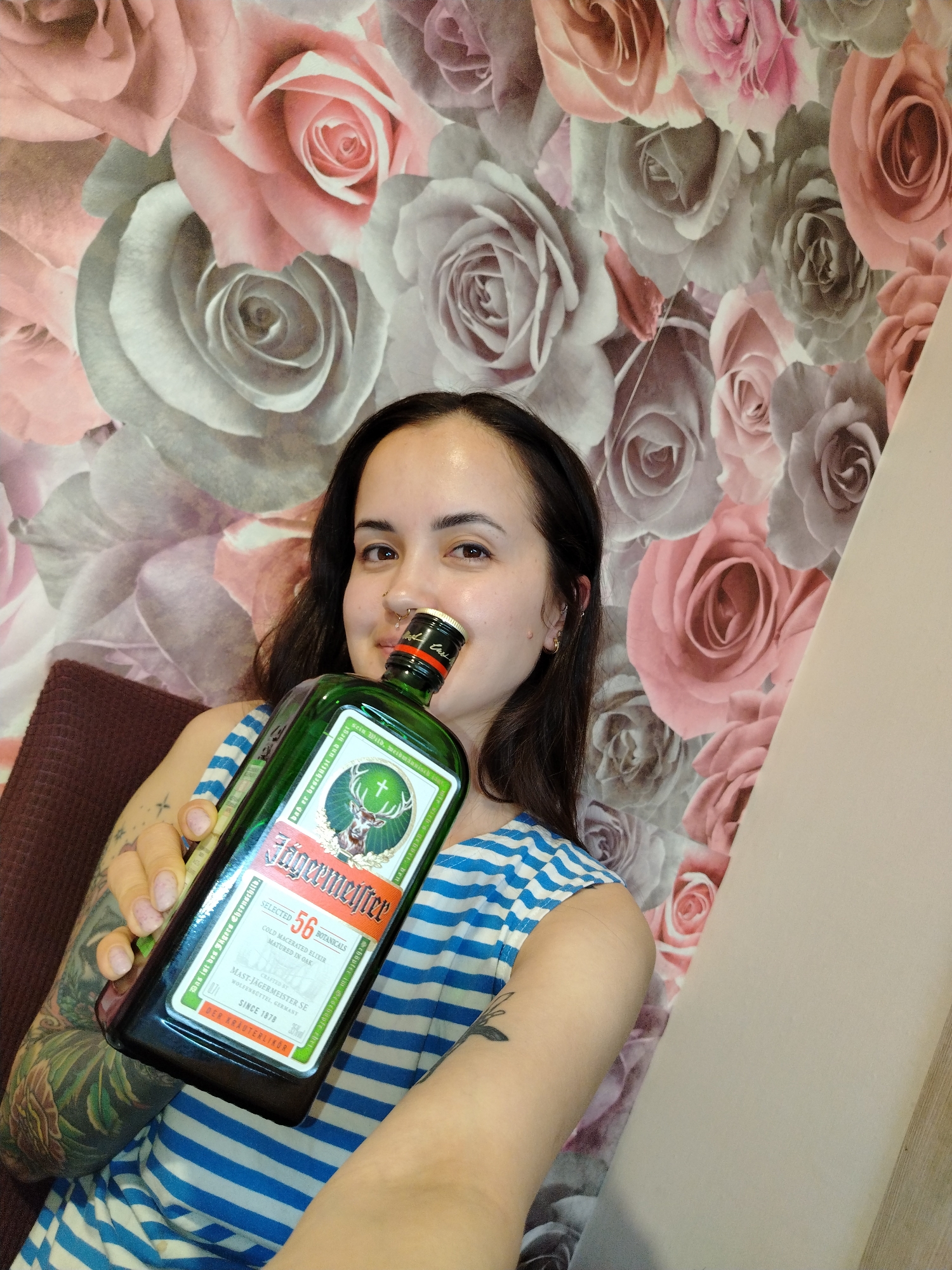 A young woman with dark hair and a nose ring is holding a bottle of Jägermeister up to her mouth. She is wearing a blue and white striped shirt and has tattoos on her left arm. The background is a wall with a floral pattern of pink and gray roses. There are no other specific details available to determine the location as Noviy, Russia.