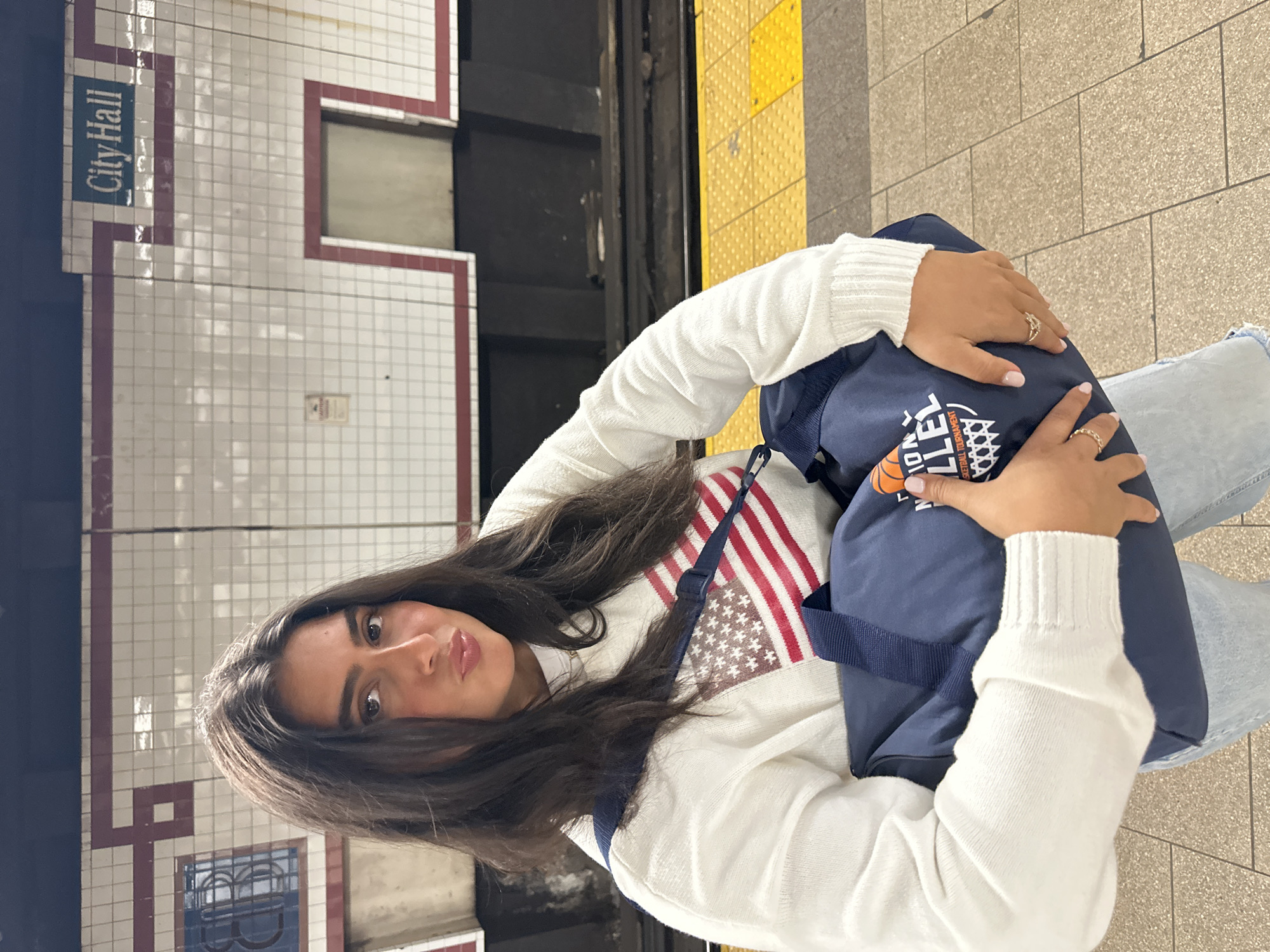 A woman is sitting at the City Hall subway station in New York City. She has long, dark hair, and she is wearing a white sweater with a faded American flag on it. She is holding a blue bag with the word “basketball” on it. The subway station wall behind her is white tiles with red and blue trim. The platform edge is lined with yellow tactile paving.