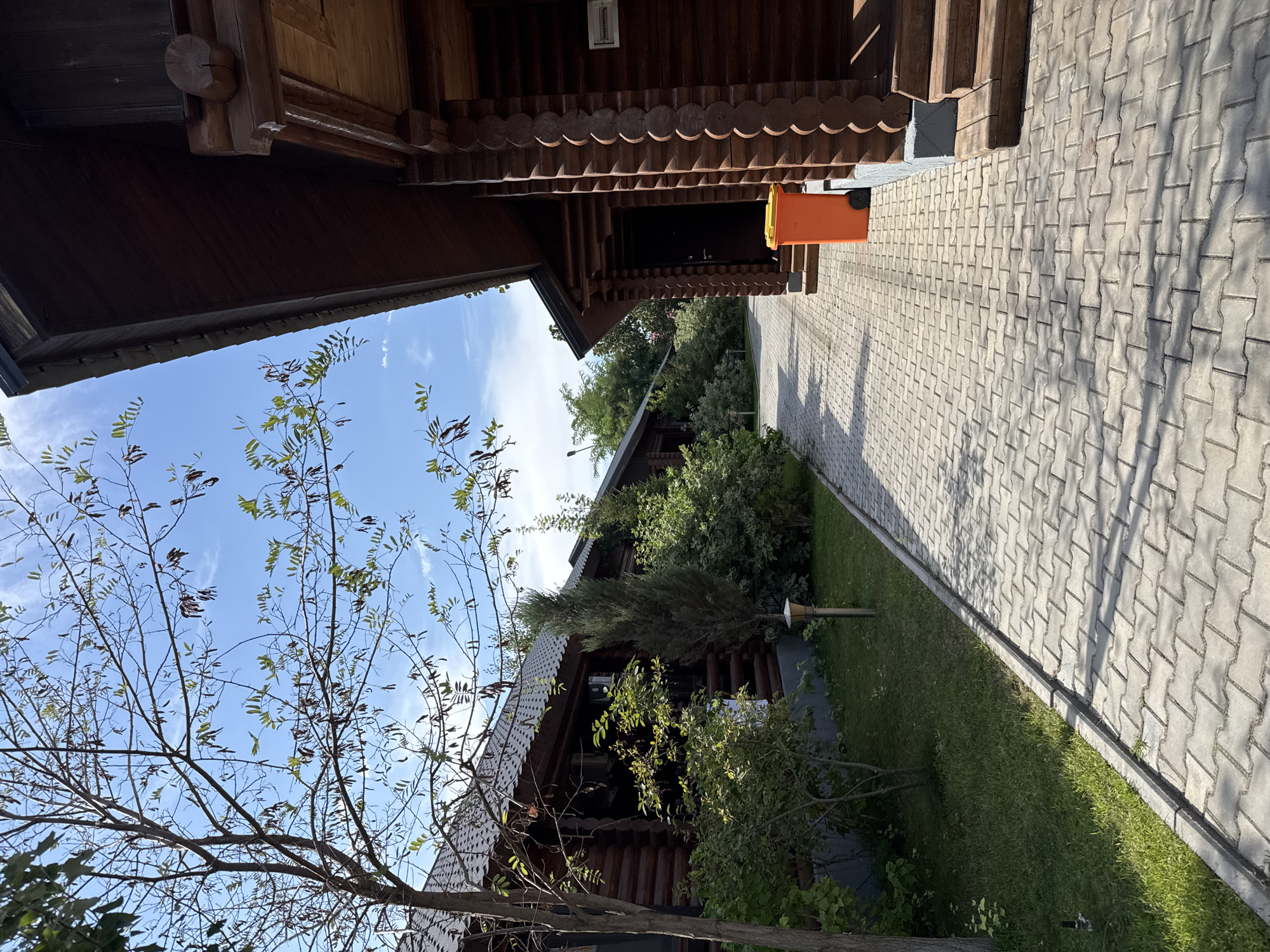 The image depicts a pathway constructed from interlocking concrete pavers, leading through a well-maintained outdoor area. To the left of the pathway, a manicured lawn is bordered by various shrubs and a slender tree with sparse foliage and seed pods. Further back on the left, a long, single-story building with a dark, patterned roof and walls constructed from dark wooden logs is visible. This building appears to be a restaurant or similar establishment. On the right side of the pathway, a portion of another log building, also with a dark roof and prominent wooden log construction, is prominently featured. This building has a covered entrance with wooden steps and an orange trash receptacle positioned near the entrance. The background consists of lush green trees and a bright blue sky with scattered white clouds. The overall scene suggests a pleasant, possibly resort-like, environment. The image is tagged with the location of Ақши, Kazakhstan, implying this is a scene from that region. No people or active interactions are present in the image.