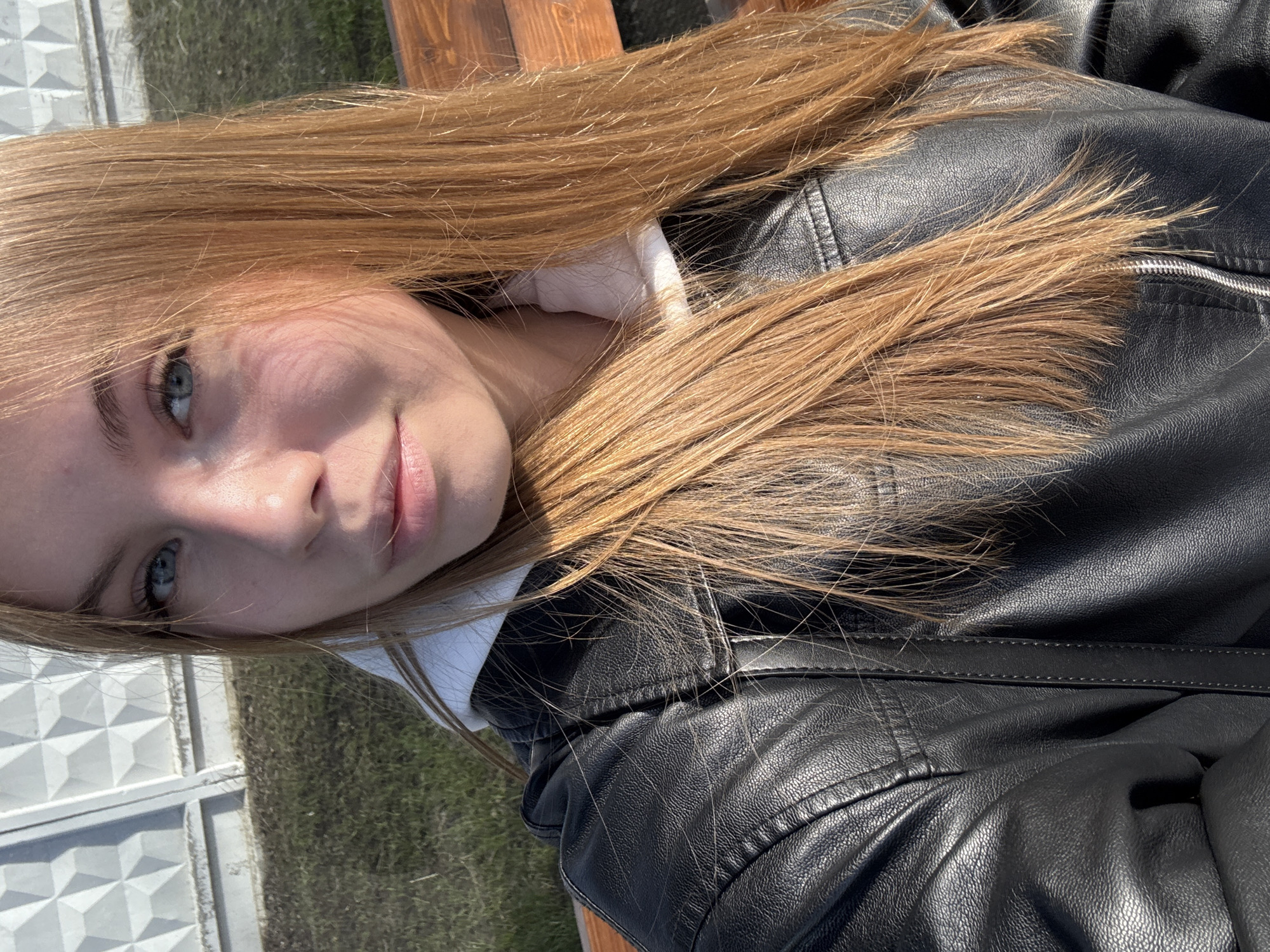 A young woman with light skin, blue eyes, and long blonde hair is facing the camera. She is wearing a black leather jacket and a white hoodie. She is sitting on a wooden bench. In the background, there is a white fence and green grass. The location is likely in the city of Chaikovsky, Russia.
