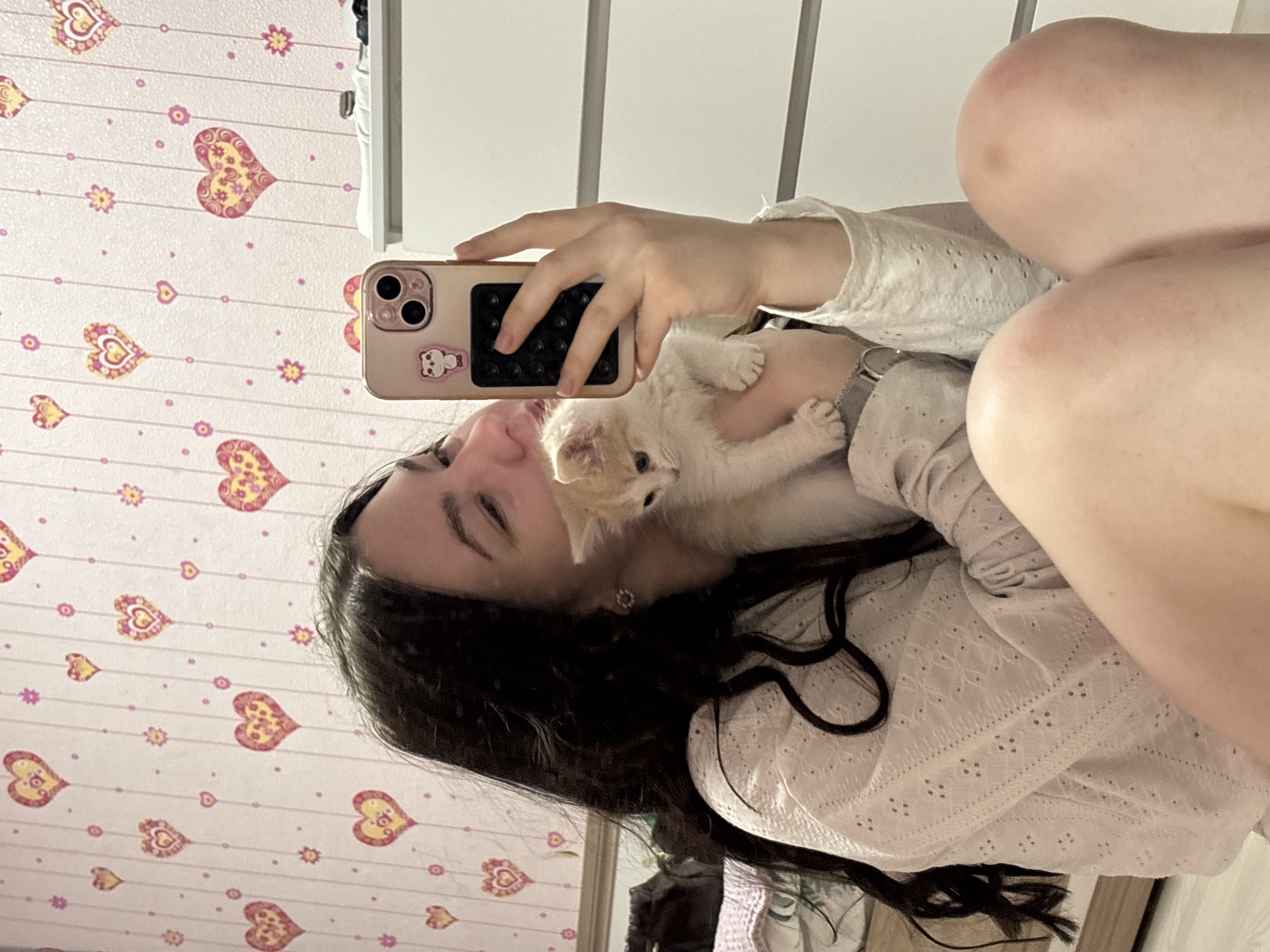 The image is a mirror selfie showing a person holding a small, fluffy kitten. The person, with long, dark hair, is wearing a light-colored, long-sleeved top with intricate embroidered or eyelet details. Their face is visible, with a gentle expression and a soft smile, looking towards the kitten or the camera.
A very young, light-colored kitten, possibly cream or pale ginger, is nestled securely in the person's arm against their chest. The kitten's tiny face and paws are visible, and it appears calm and comfortable.
The person is holding a light-colored smartphone, likely rose gold or gold, with a triple-lens camera system on the back. The phone has a light-colored case adorned with a small sticker of a cartoon bear or similar animal.
The background is a wall covered in wallpaper featuring a playful design. The pattern includes horizontal stripes with small dots, interspersed with larger heart shapes decorated with yellow floral patterns, and smaller purple floral or star-like elements. Parts of a white piece of furniture or wall paneling are visible in the reflection, indicating an indoor setting where the selfie was taken using a mirror. The overall mood conveyed is one of warmth and affection. No discernible text is visible.