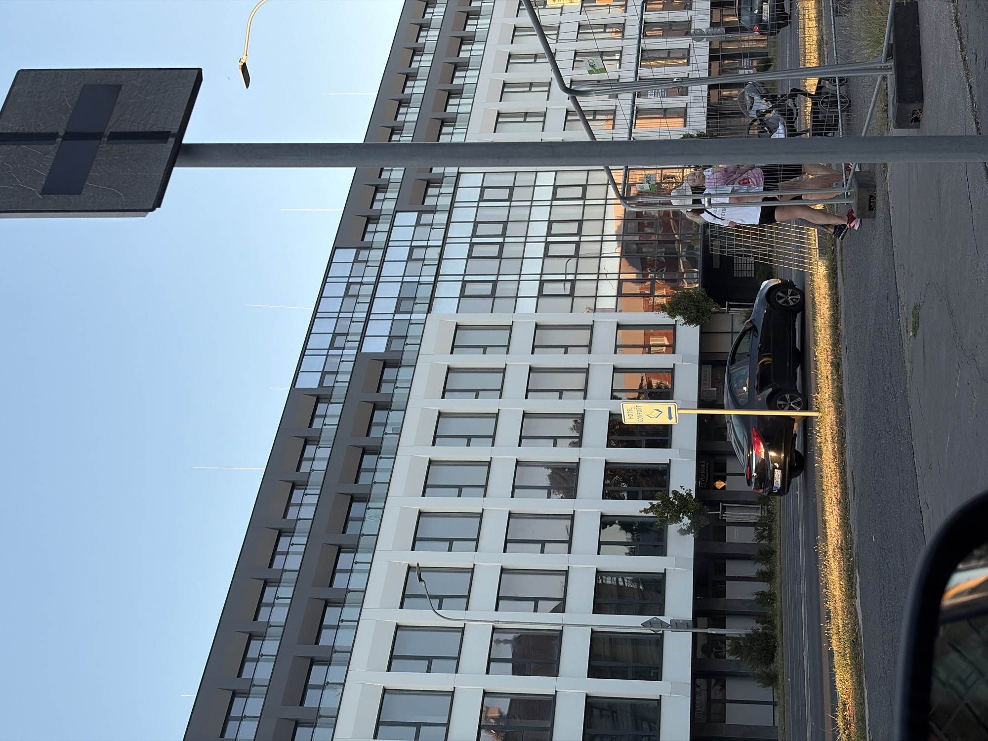 The image shows a modern building with a glass facade in the city of Nitra, Slovakia. The building appears to be an apartment complex or office building, with multiple stories and balconies. In front of the building, there is a street with a black car driving on it. There are also some pedestrians walking on the sidewalk, and a stroller. The scene is illuminated by the warm light of the setting sun, casting long shadows across the street. The sky is clear and blue, indicating good weather. There is a traffic sign pole in the foreground, and a wire fence in the middle ground. A yellow sign with the text "HOTEL COMFORT" and an arrow pointing to the left is visible on a pole. The overall impression is one of urban life during a pleasant evening.