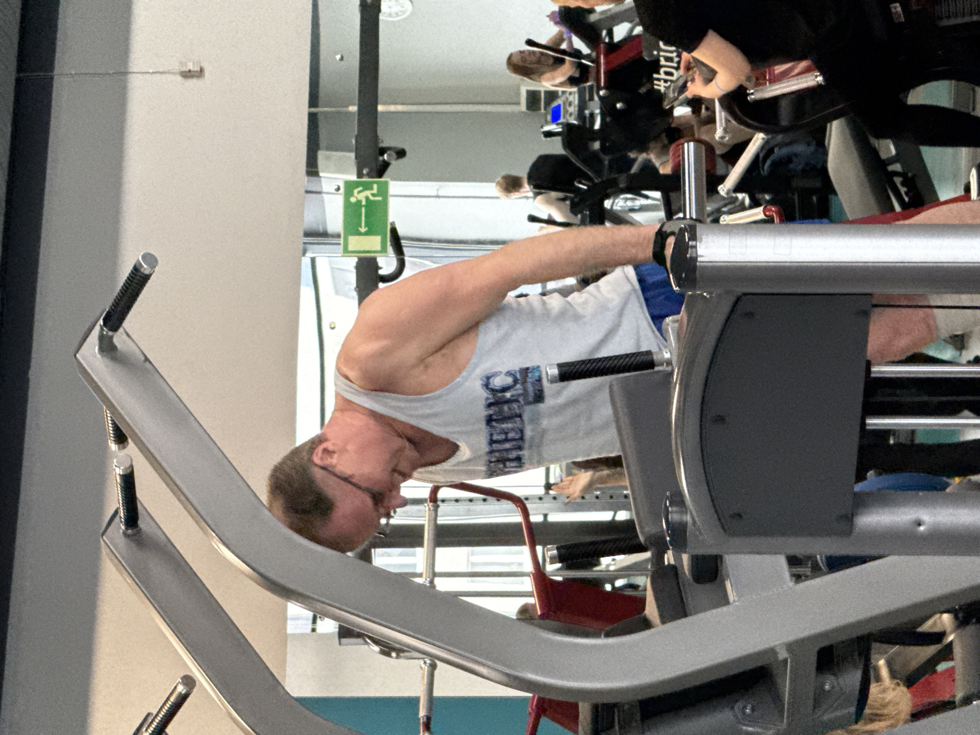 The image shows a man in a gym using a weight machine. He is wearing a light gray tank top and blue shorts. The gym appears to be modern, with other people visible in the background using exercise equipment. The location is difficult to determine from the image alone and a location in Yekaterinburg, Russia cannot be definitively confirmed.