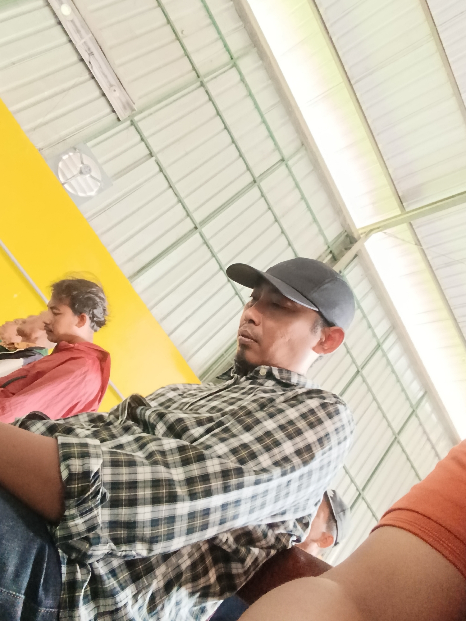The image captures three individuals sitting indoors, possibly in a communal area or shelter, in Kawunganten Lor, Indonesia. The man in the foreground, wearing a grey baseball cap and a plaid shirt, is partially visible, with his face turned away from the camera, and he appears to be looking down. To his left, another man in a red shirt is seen from the side, also looking downwards, with a mustache and dark hair. Further to the left, a third person is barely visible, wearing a light-colored garment. The environment features a distinctive yellow wall on the left and a corrugated metal ceiling with visible beams overhead. The lighting suggests an indoor setting, potentially during daylight hours.