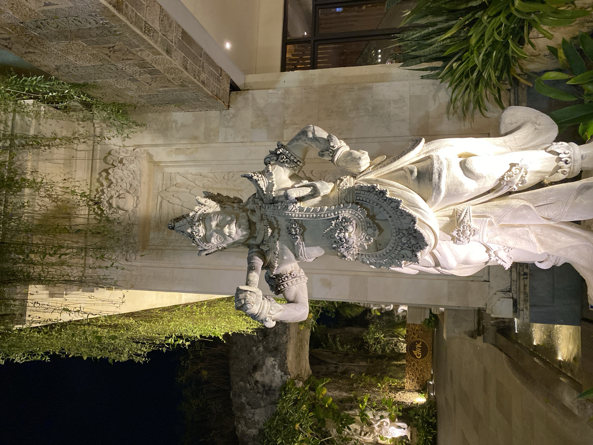The image shows a nighttime exterior scene in Kuta Selatan, Indonesia. A large, stone statue of a warrior with ornate headdress and clothing stands in the foreground, illuminated by spotlights. The statue is positioned in front of a light-colored stone wall with intricate carvings. Hanging plants cascade down from above, partially obscuring the wall and the statue. To the left of the statue, a small fountain with cascading water is visible, and beyond that, a pathway and more greenery. In the background, a sign with "GWK" is illuminated. To the right of the statue, a large window with reflections of interior lights is visible, and a potted snake plant is situated in front of it. The overall ambiance is one of a beautifully lit cultural or resort setting.