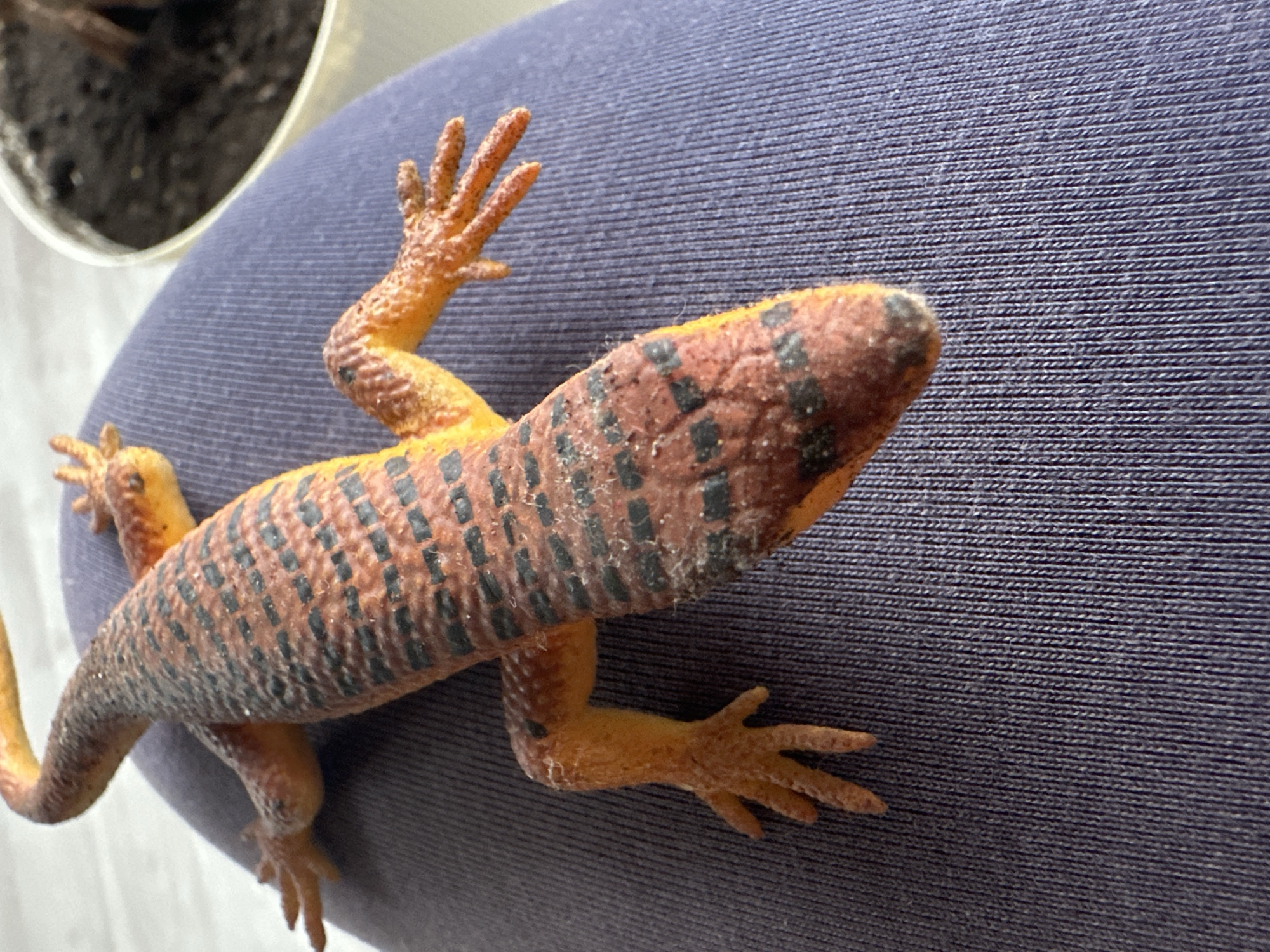 A toy lizard, primarily orange and brown with black stripes, rests on a dark blue textured surface, possibly fabric. The lizard is positioned lengthwise, with its head pointing towards the bottom right of the frame. Its limbs are splayed out. In the upper right corner, a portion of a white pot containing dark soil is visible. The scene appears to be indoors, with neutral lighting. There is no discernible location in the city of Чернишня, Russia.